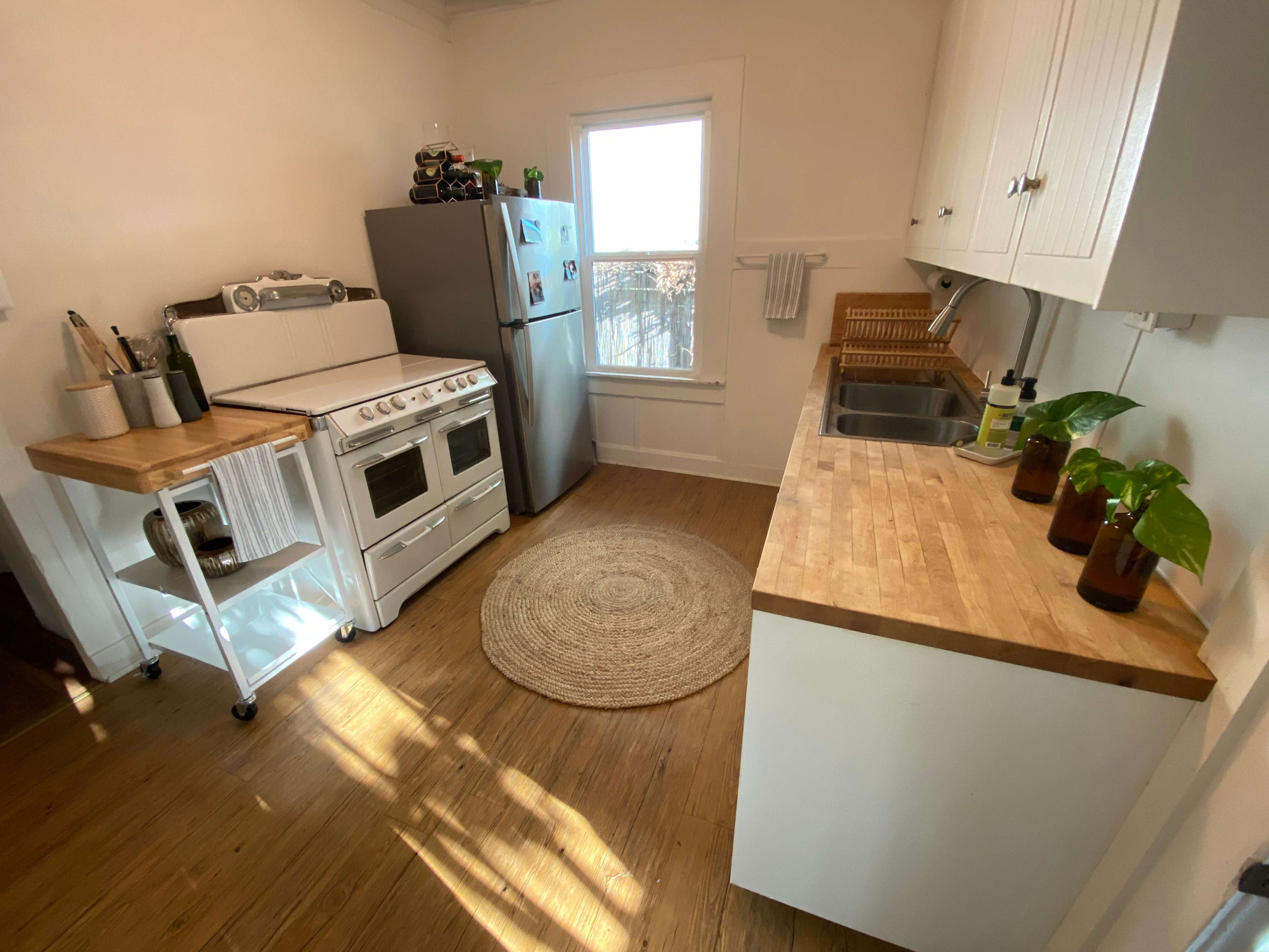 The kitchen features a white stove and silver refrigerator, with wooden countertops and a round rug on the floor, illuminated by natural light from the window.