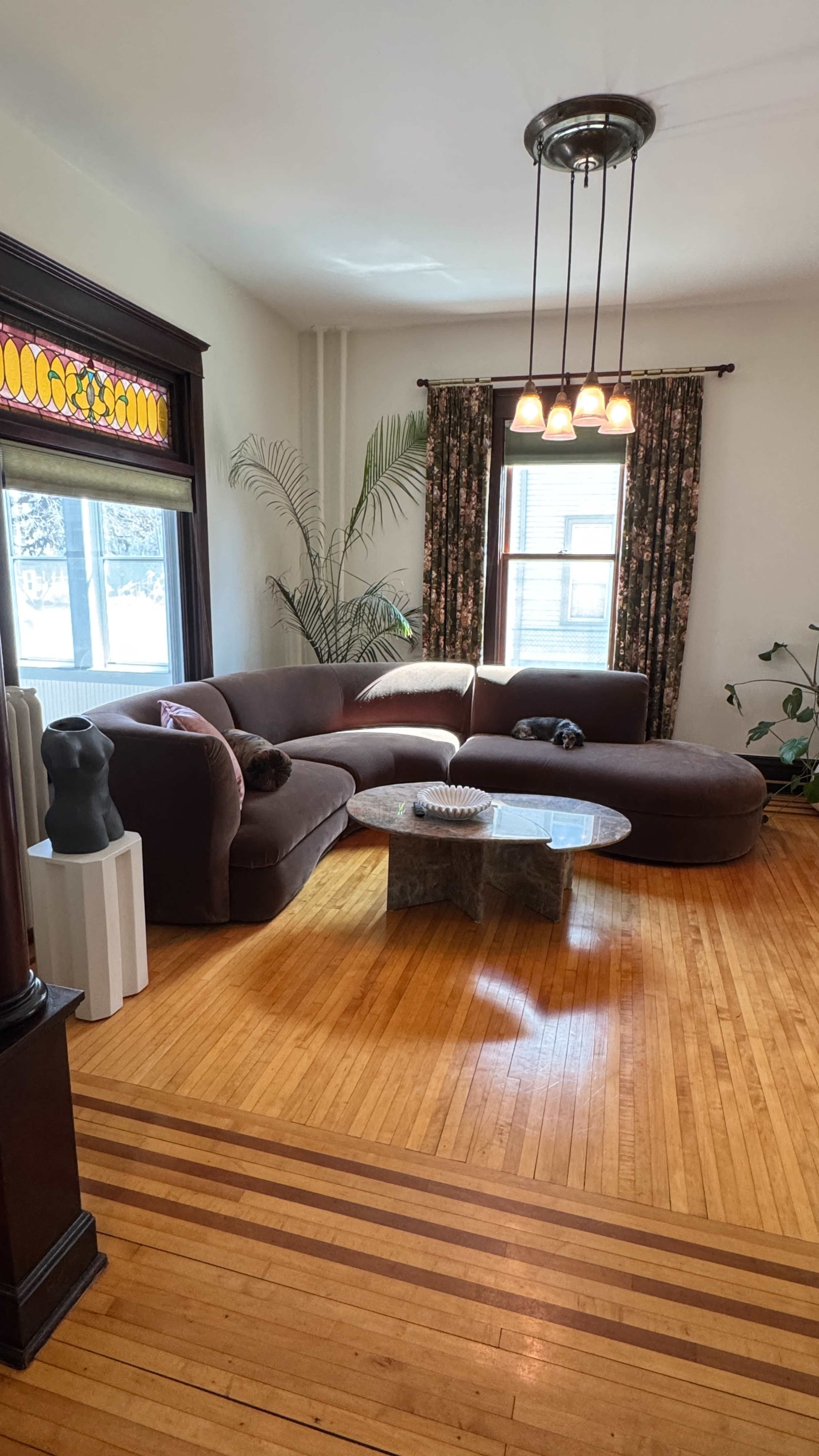 The image shows a living room featuring a large, curved brown sofa, a circular stone coffee table, and floral curtains, with a potted plant in the corner.