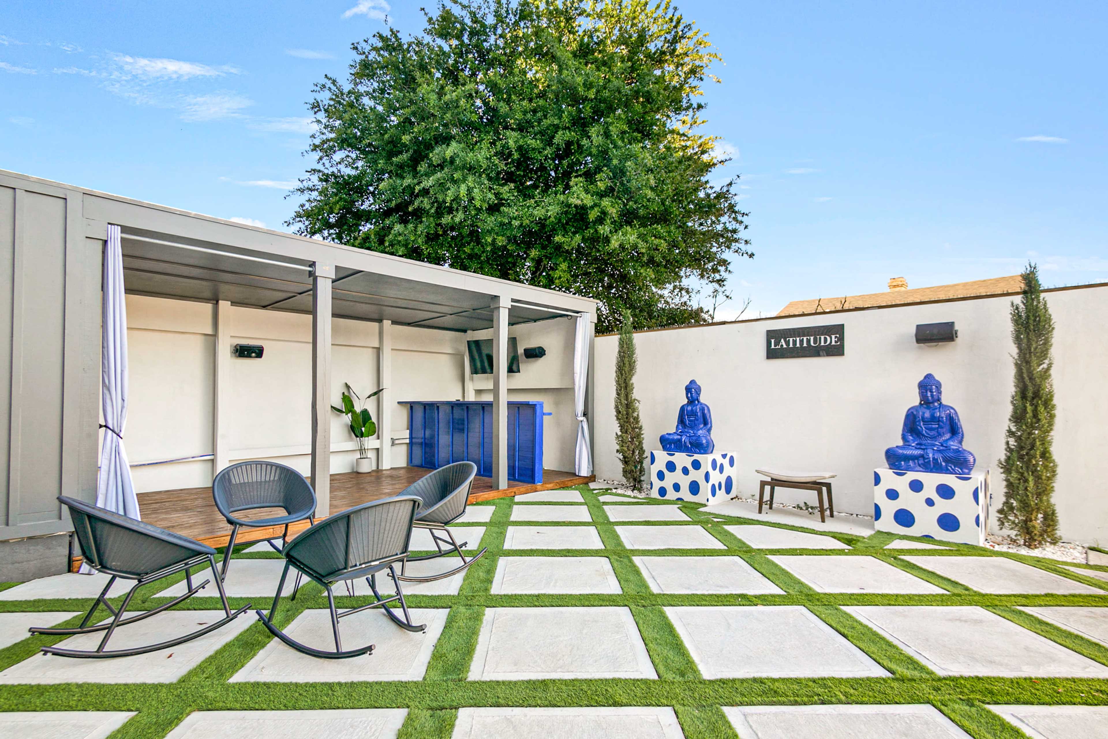 A patio area with four black lounge chairs arranged around a small table, a blue bar in the background, and two large blue Buddha statues on pedestals.