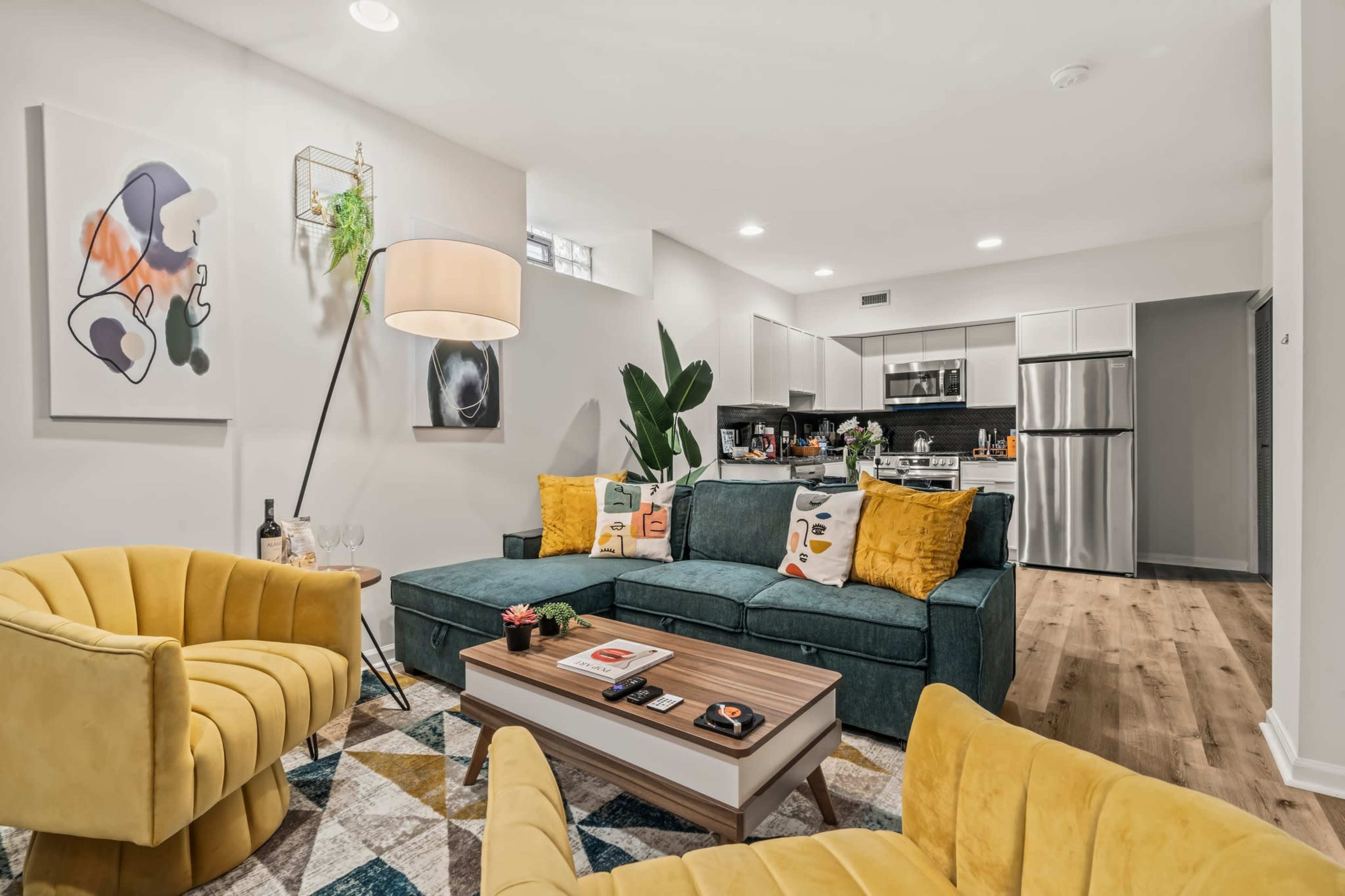 The image shows a modern living room with a green sofa, yellow accent chairs, a coffee table, and a kitchen visible in the background.
