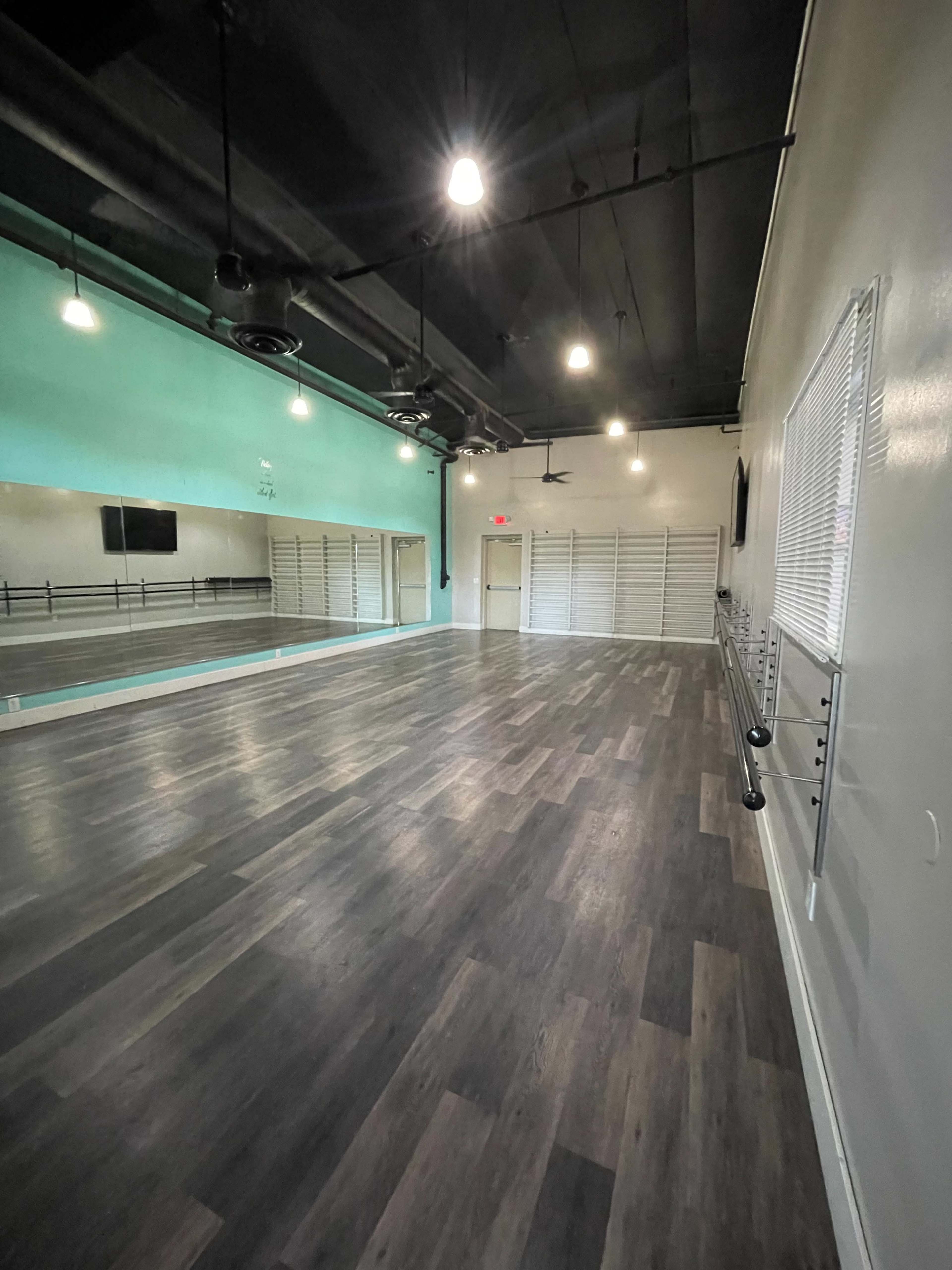 A spacious, well-lit dance studio with wooden flooring, large mirrors, and a wall of storage racks.