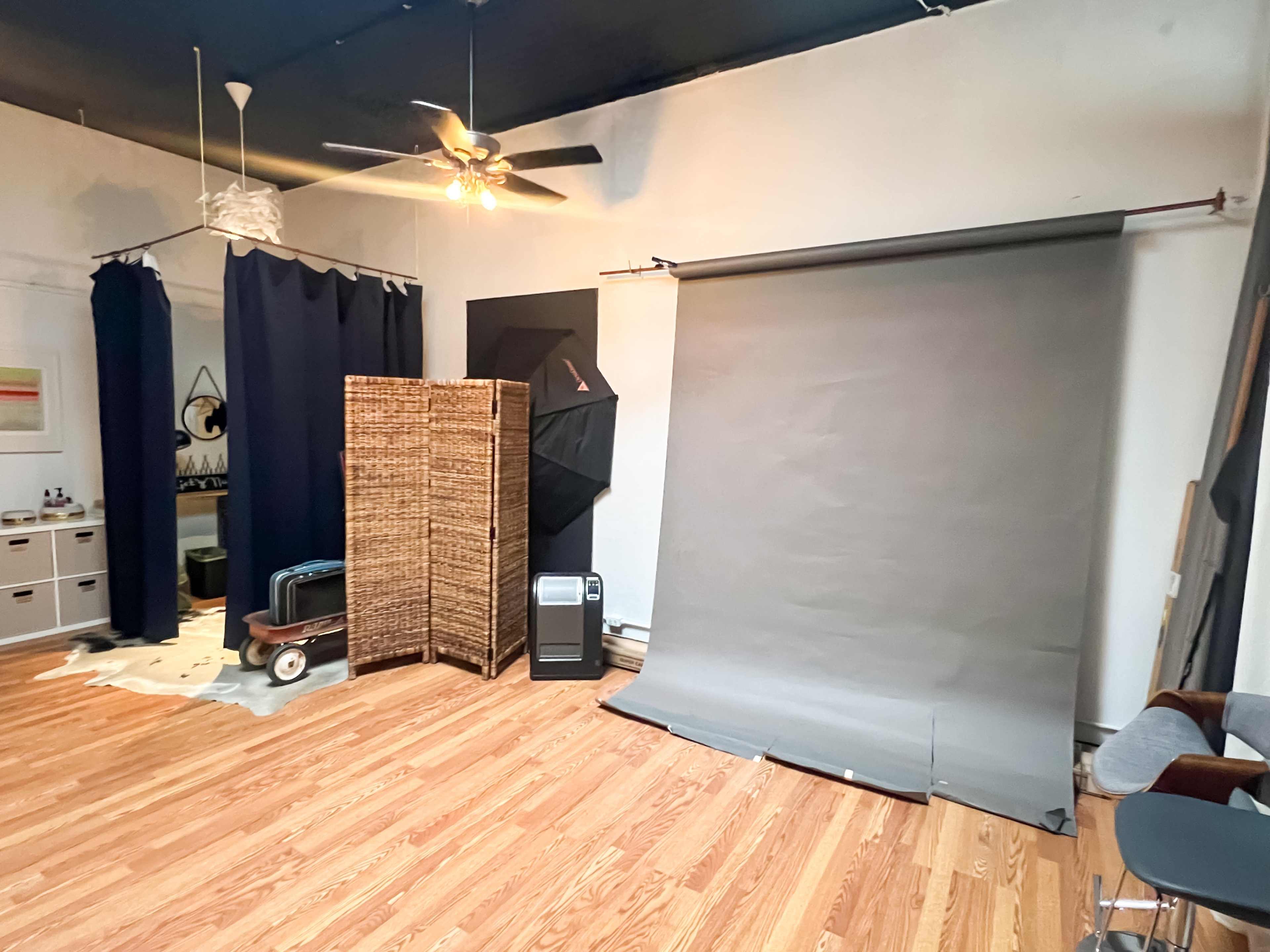 A spacious room with wooden flooring, featuring a gray backdrop, a screen divider, a fan, and various pieces of furniture.