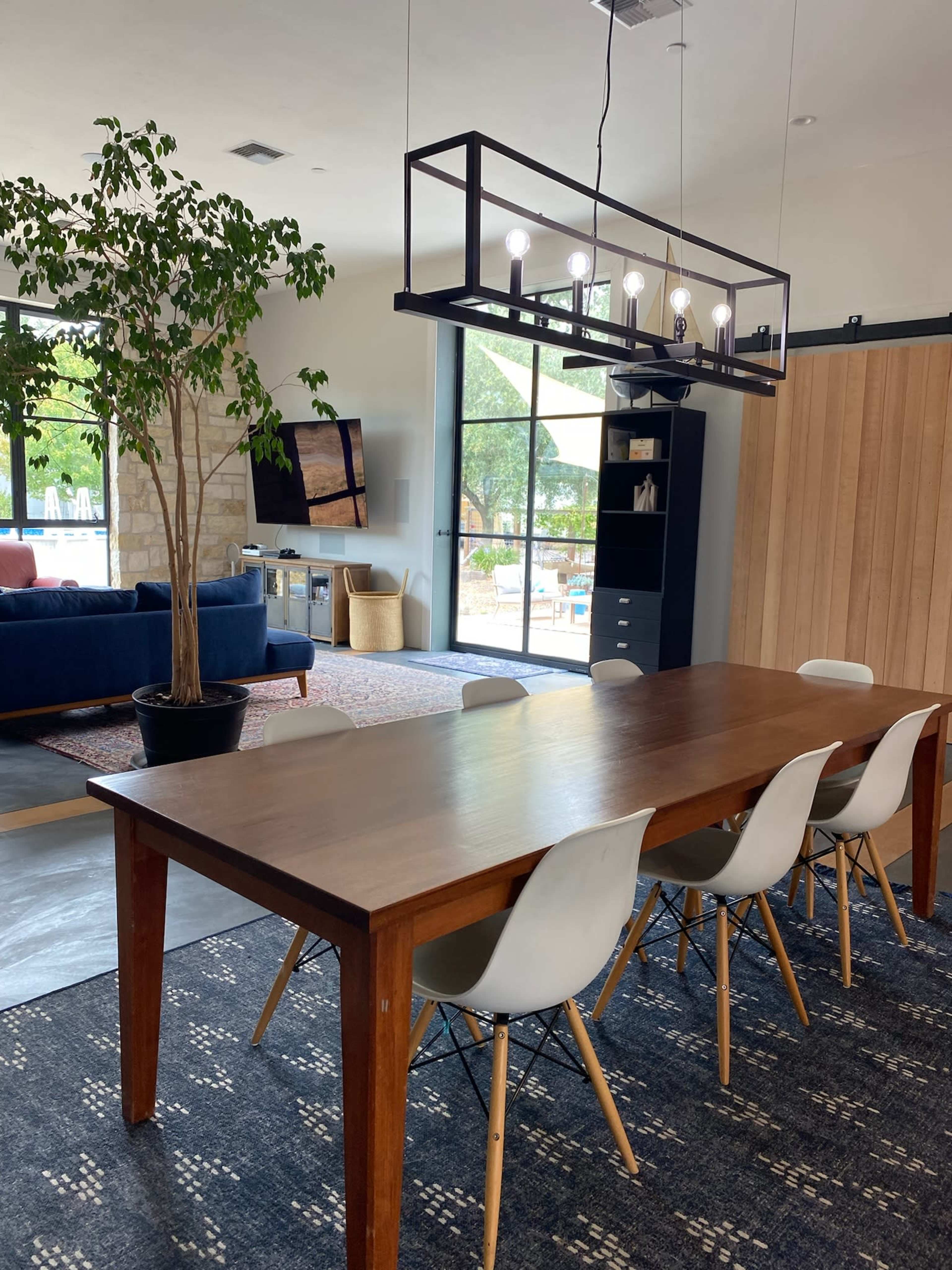 A wooden dining table is set with white chairs beneath a modern light fixture in a spacious room featuring large windows, a television, and a potted plant.