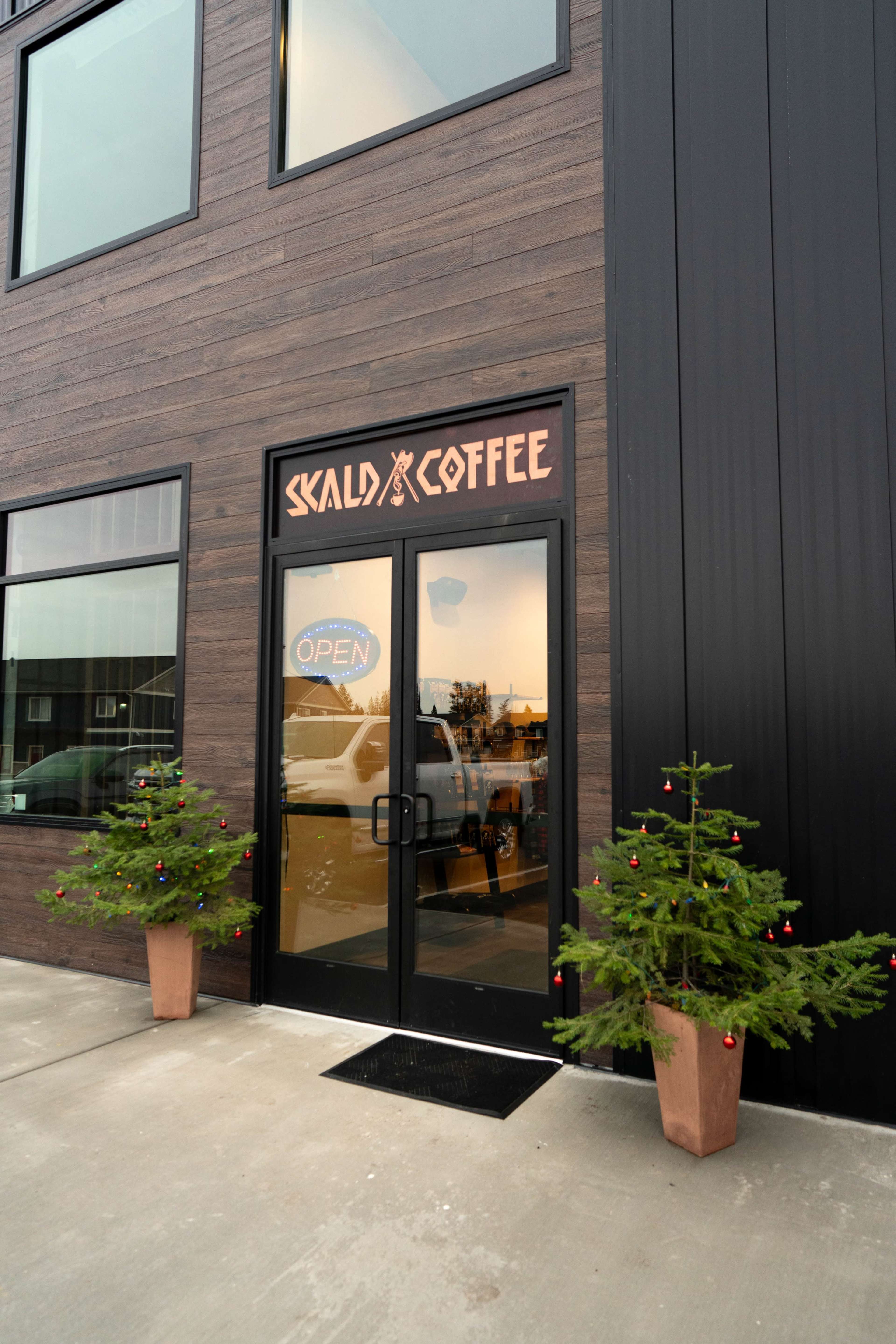 The entrance of a coffee shop features double glass doors with a sign reading "OPEN" and decorative potted trees on either side.