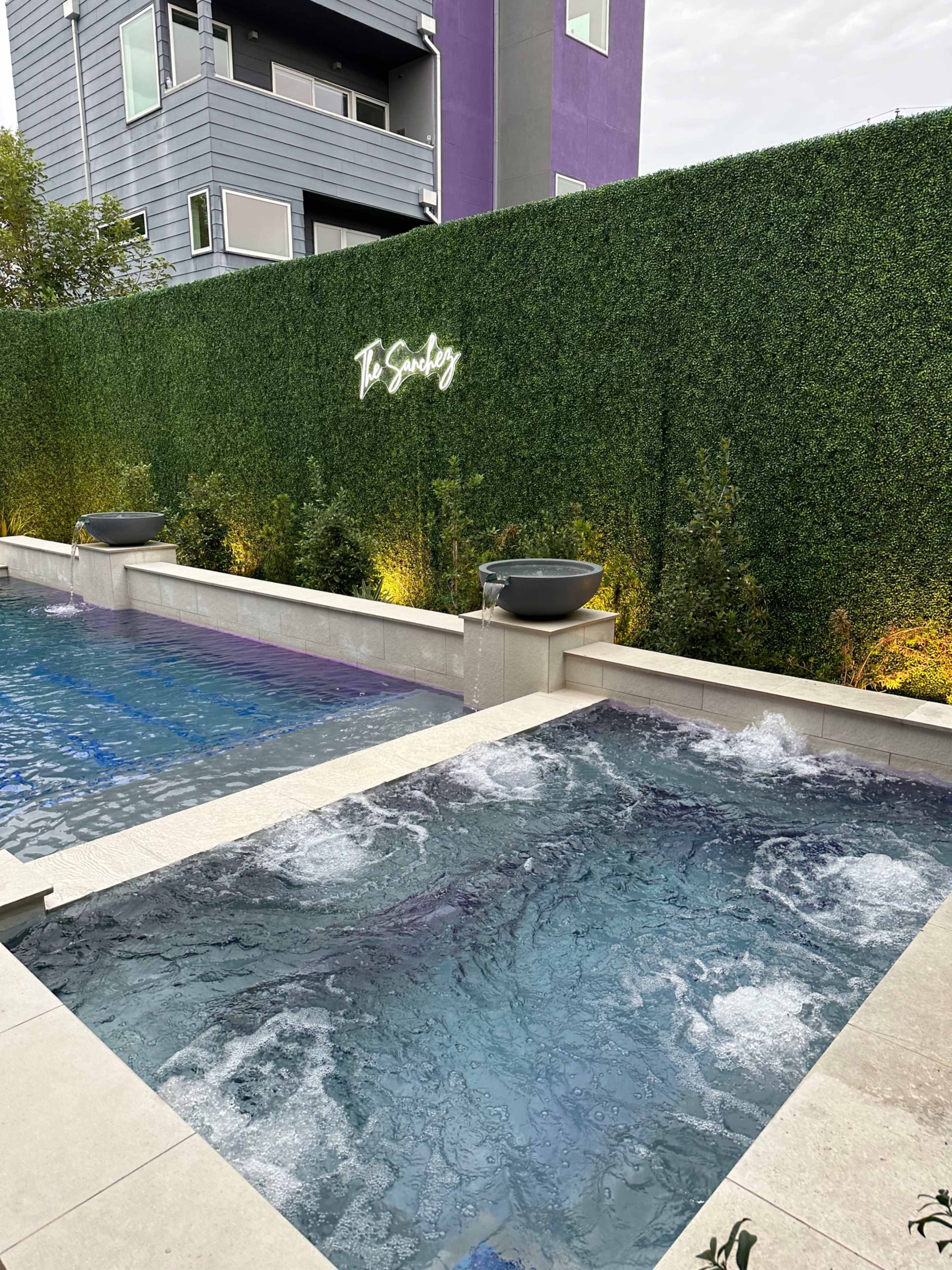 A sleek outdoor area featuring a swimming pool and a spa, surrounded by a tall green privacy hedge and illuminated by wall-mounted lights.