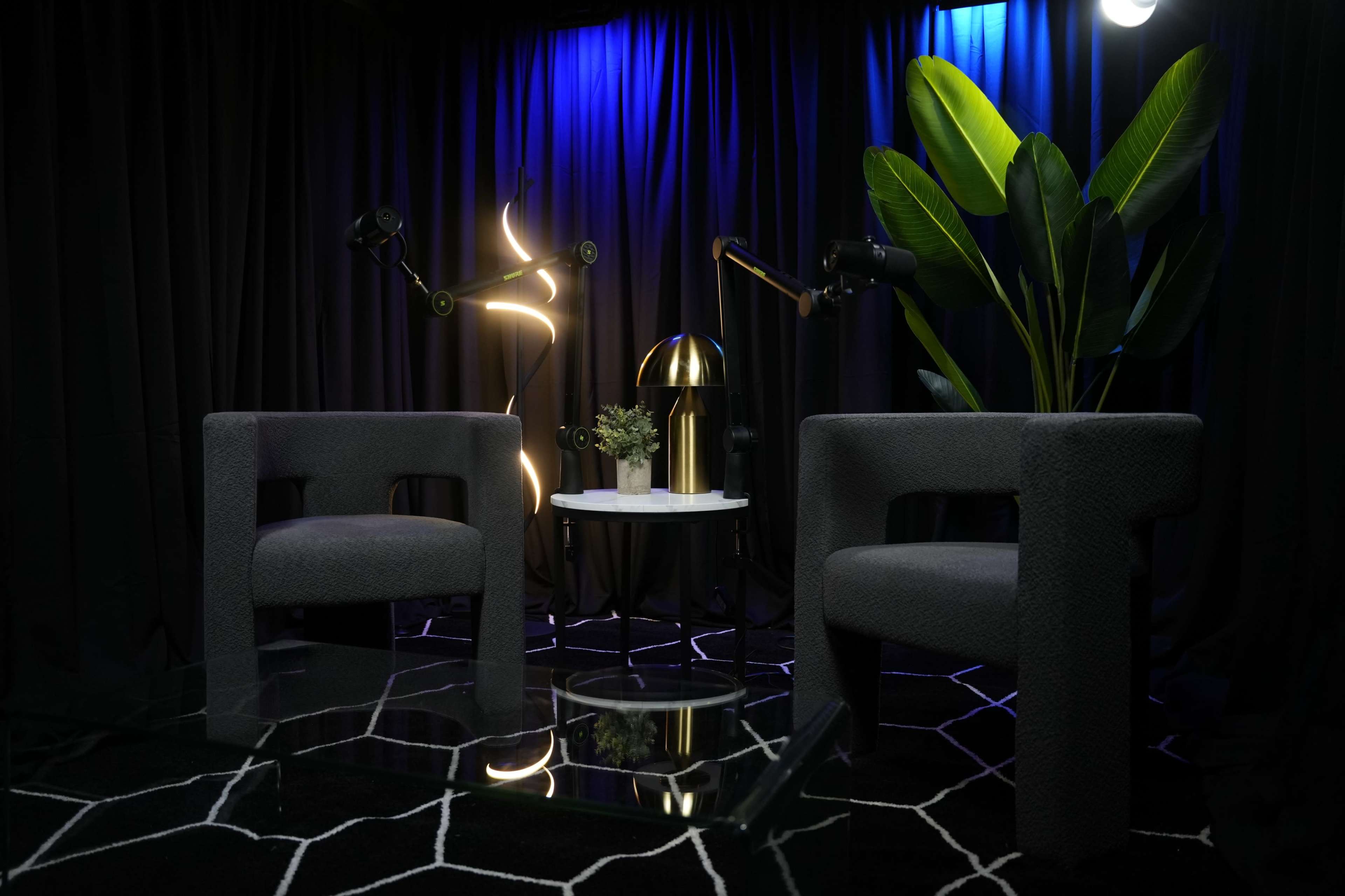 The scene features two gray chairs facing a round table, adorned with a potted plant and a lamp, all set against a backdrop of dark curtains illuminated by blue and white lighting.