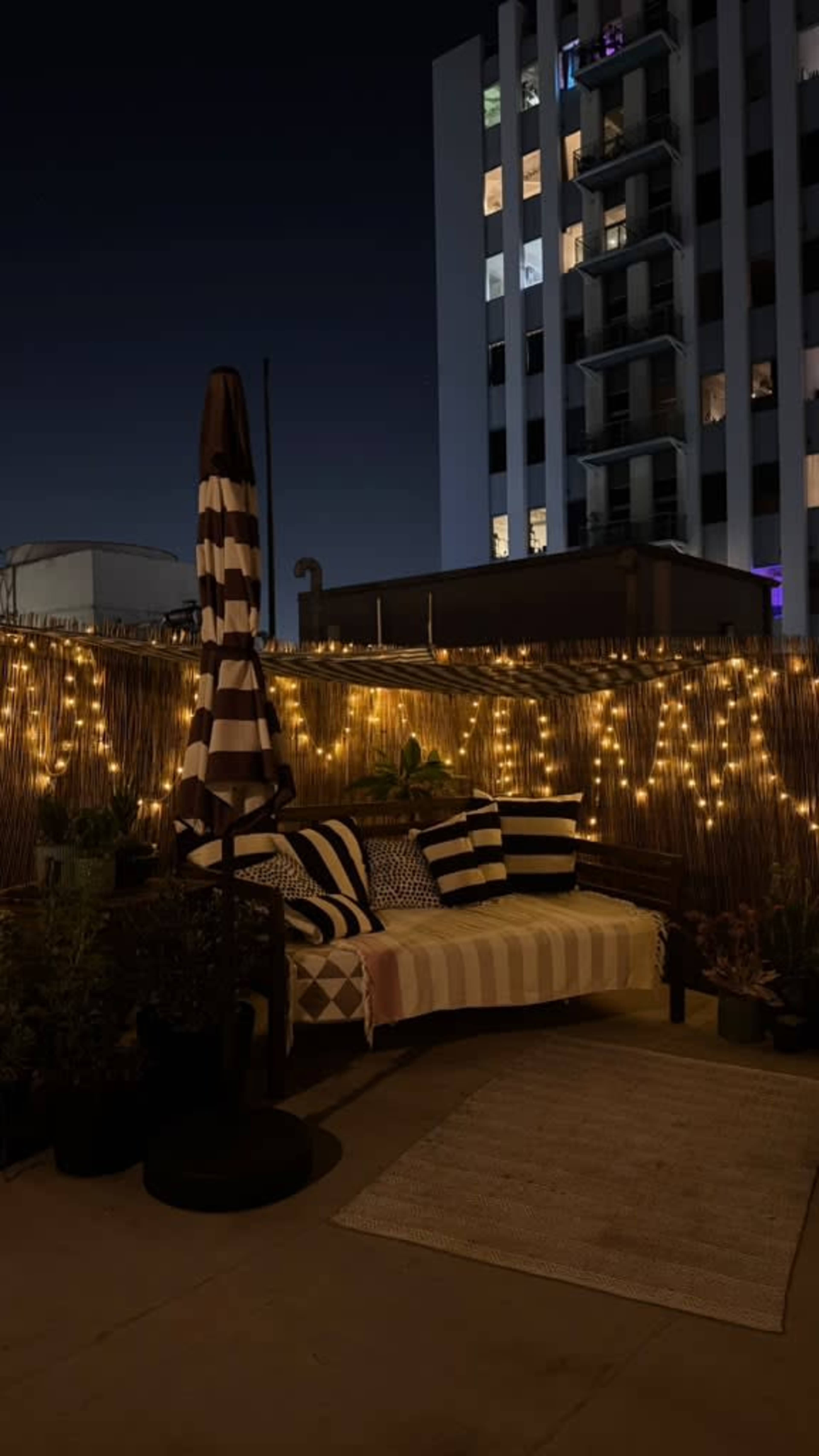 The image shows a rooftop area at night, featuring a seating space with striped cushions, a striped umbrella, string lights, and potted plants against a backdrop of a tall building.