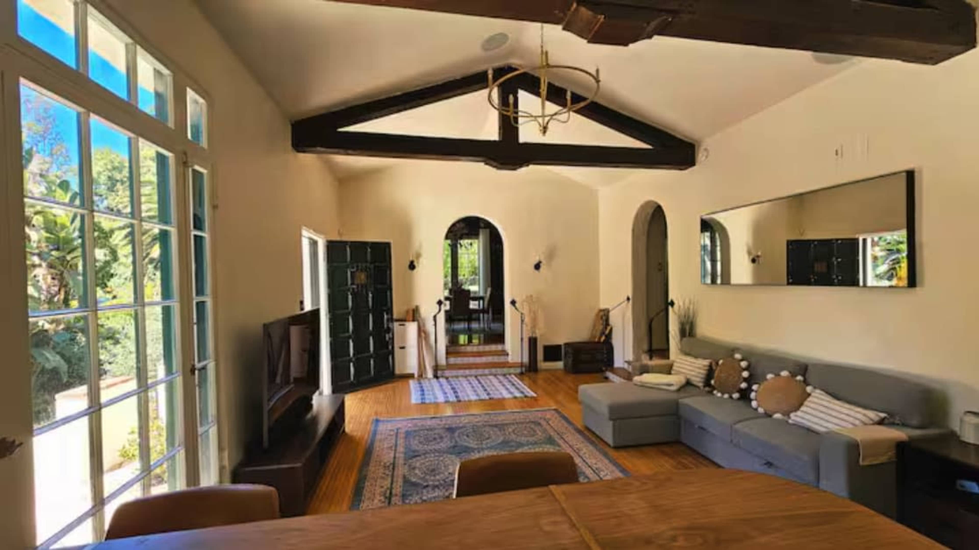 The room features a bright, open space with large windows, a sectional sofa, and wooden beams on the ceiling.