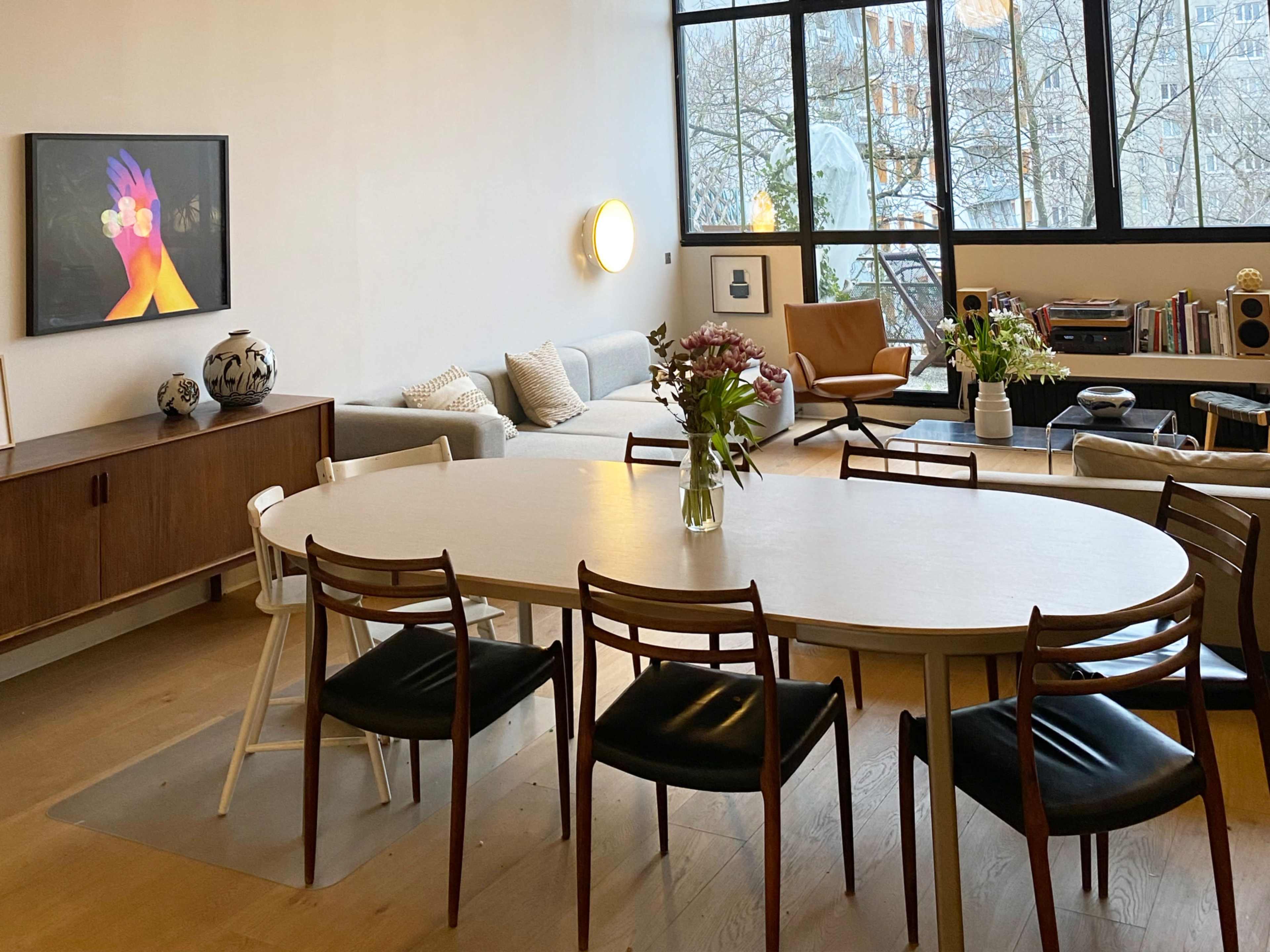 A dining area features a large oval table surrounded by wooden chairs, with a modern living space visible in the background that includes a couch and large windows.