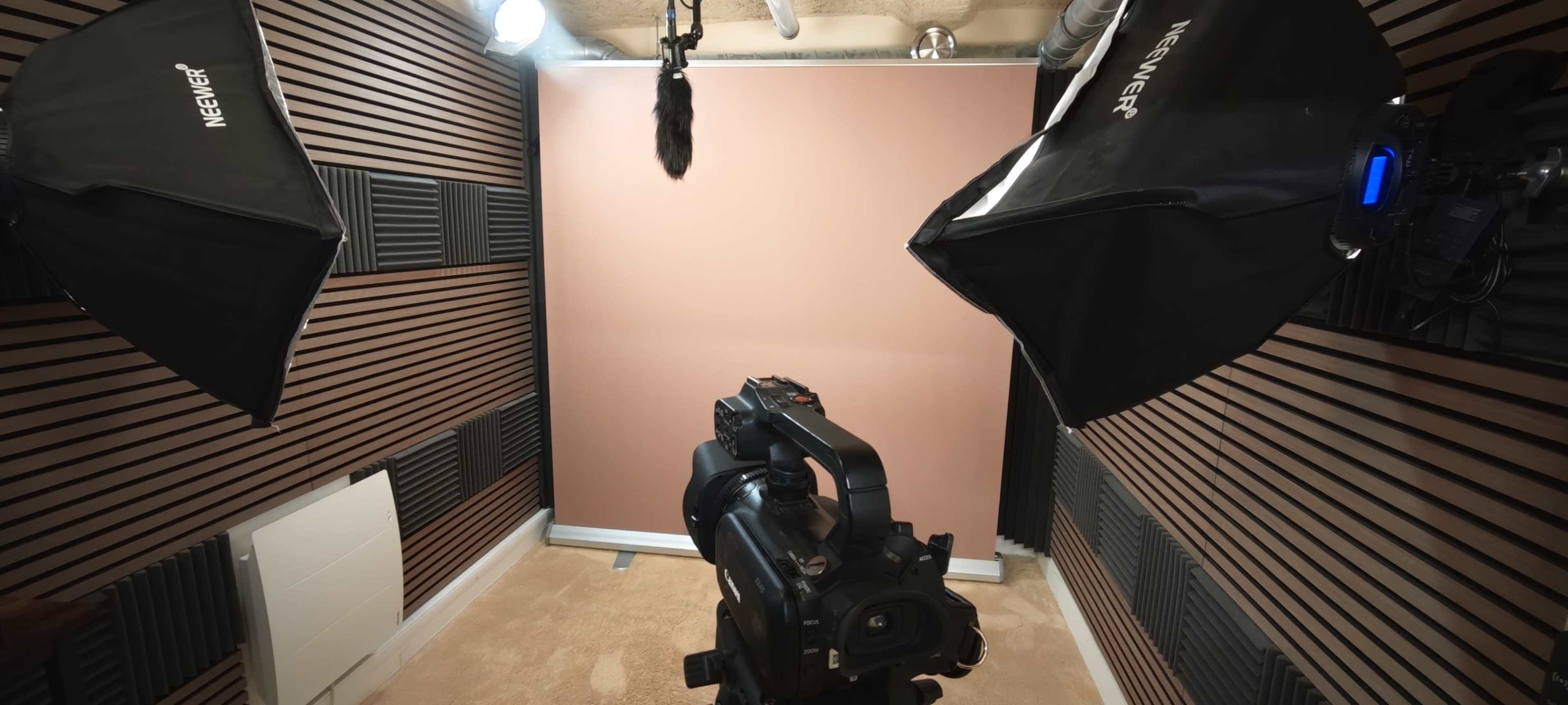 The image shows a photography setup with a camera facing a plain backdrop, flanked by softbox lights and sound equipment in a studio space.