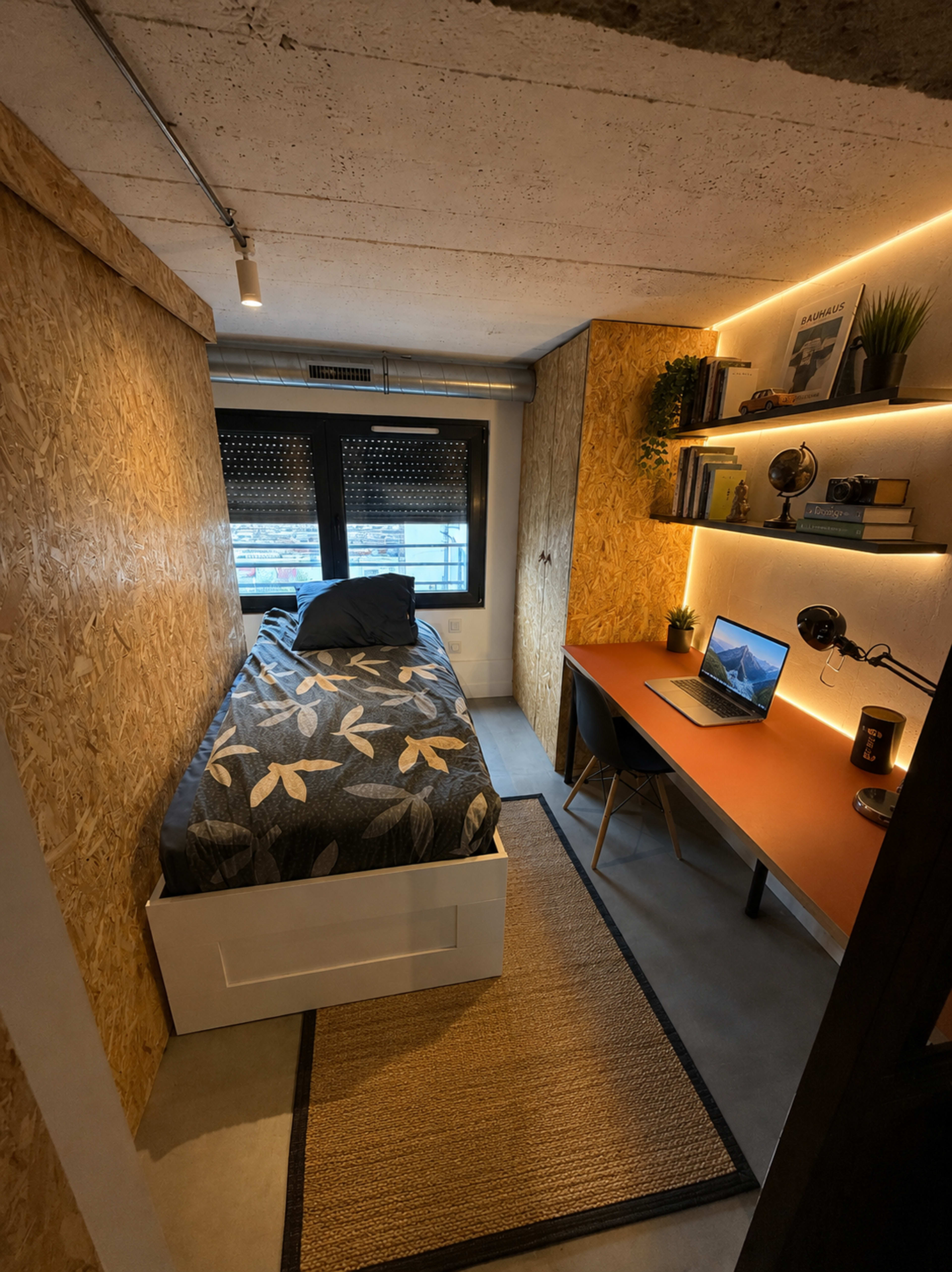 The image shows a small bedroom featuring a bed with a patterned blanket, a desk with a computer, and shelving illuminated by warm lights.