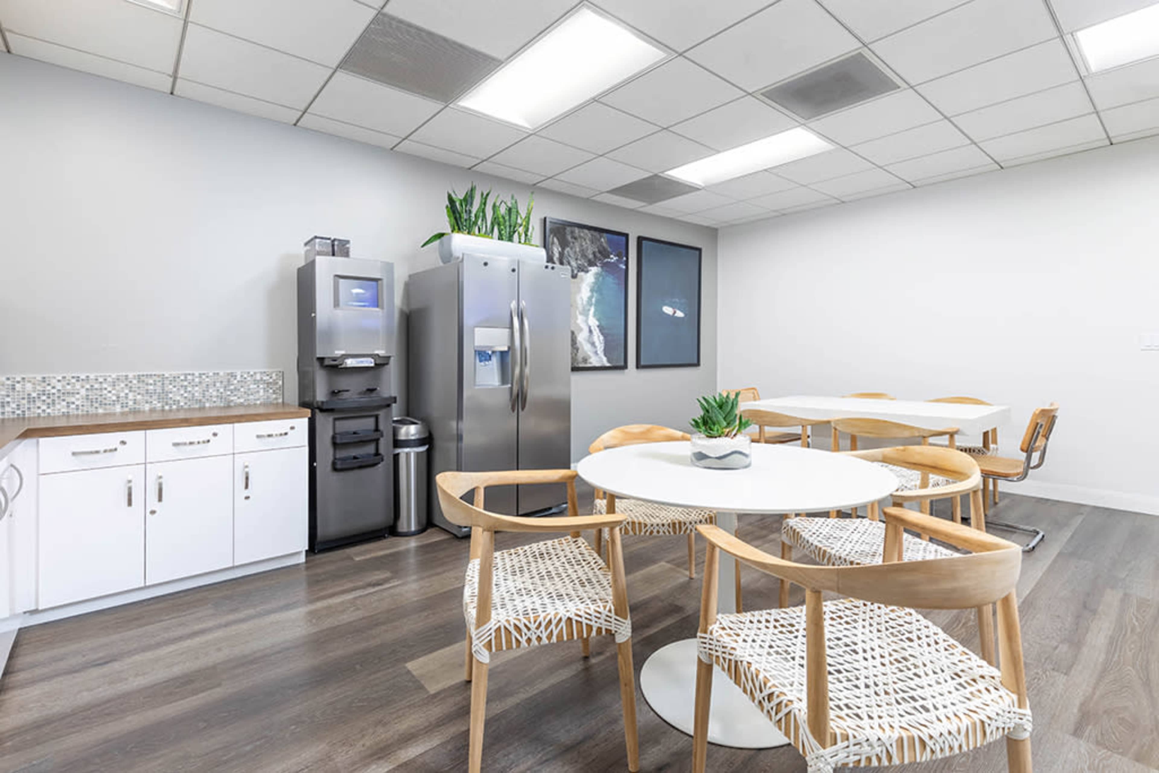 The image shows a modern break room with a round white table surrounded by wooden chairs, a refrigerator, and a kitchenette with storage cabinets.