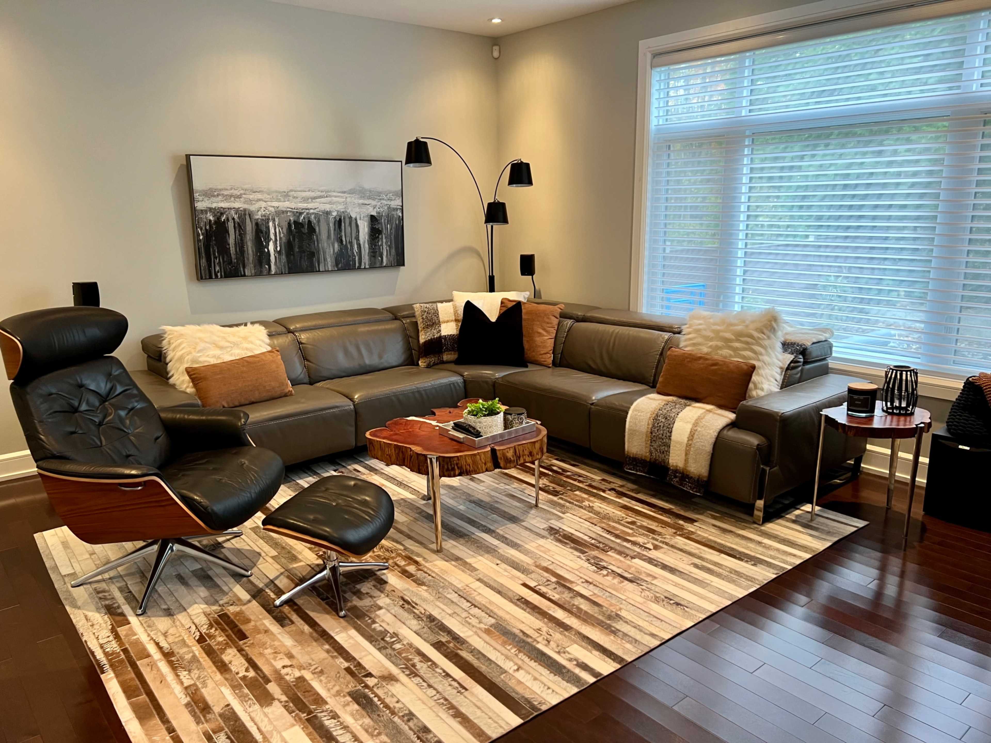 A modern living room with a large sectional sofa, a coffee table, and a lounge chair, all arranged on a patterned area rug beneath large windows with horizontal blinds.