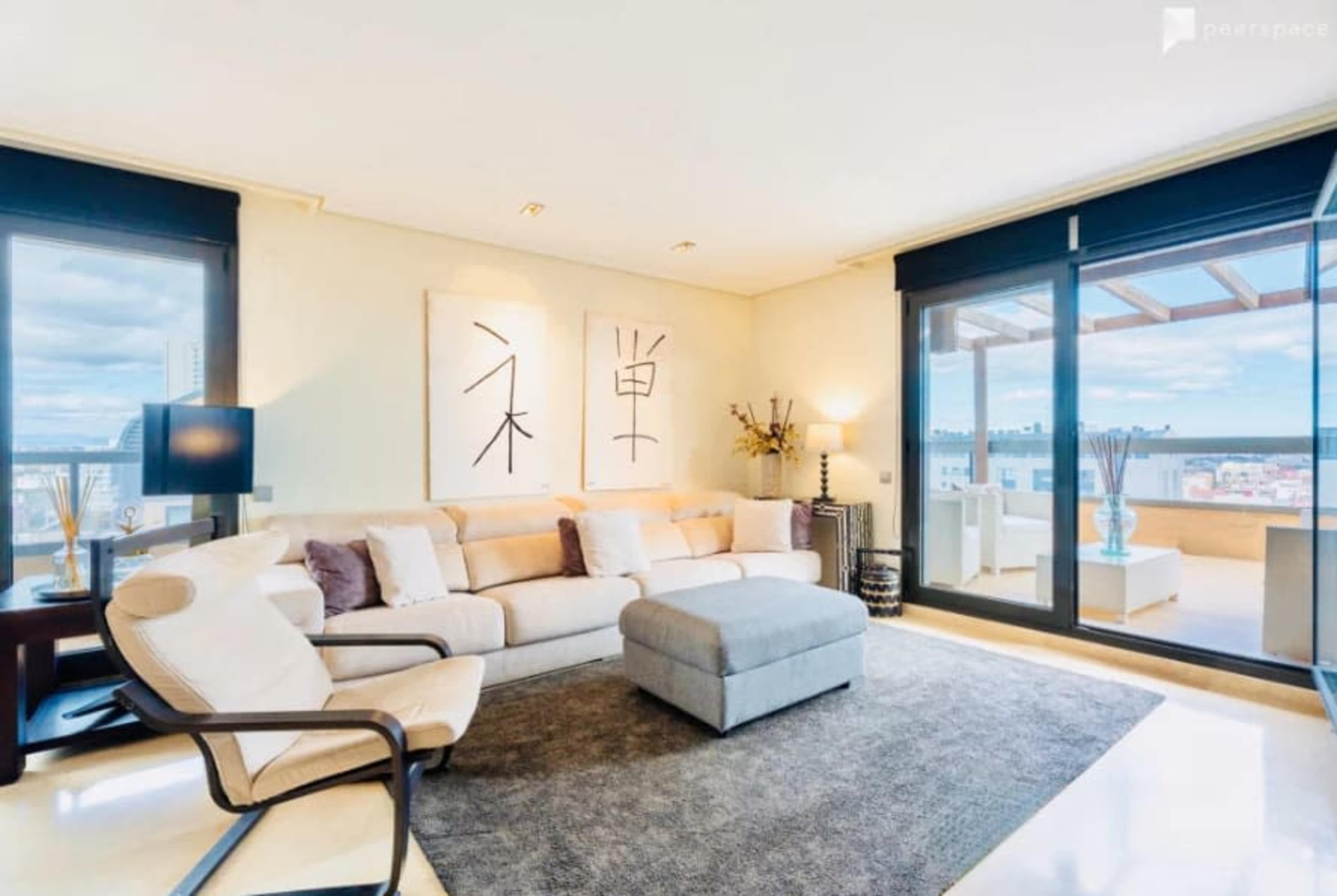 The image shows a modern living room with a beige sofa, a large ottoman, abstract wall art, and large windows that let in natural light.
