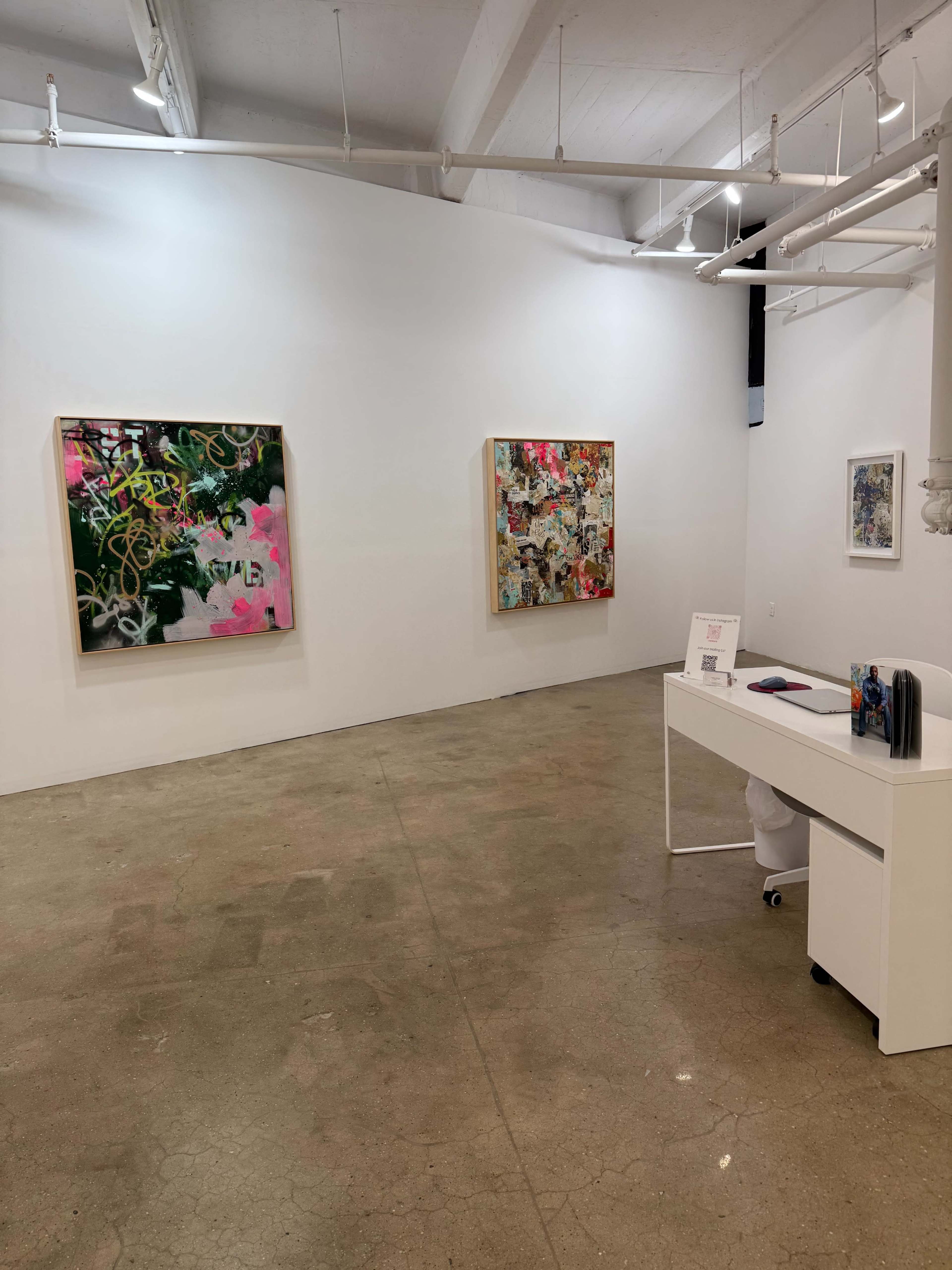 The image shows an art gallery with two colorful abstract paintings hung on white walls and a minimalist white desk in the foreground.