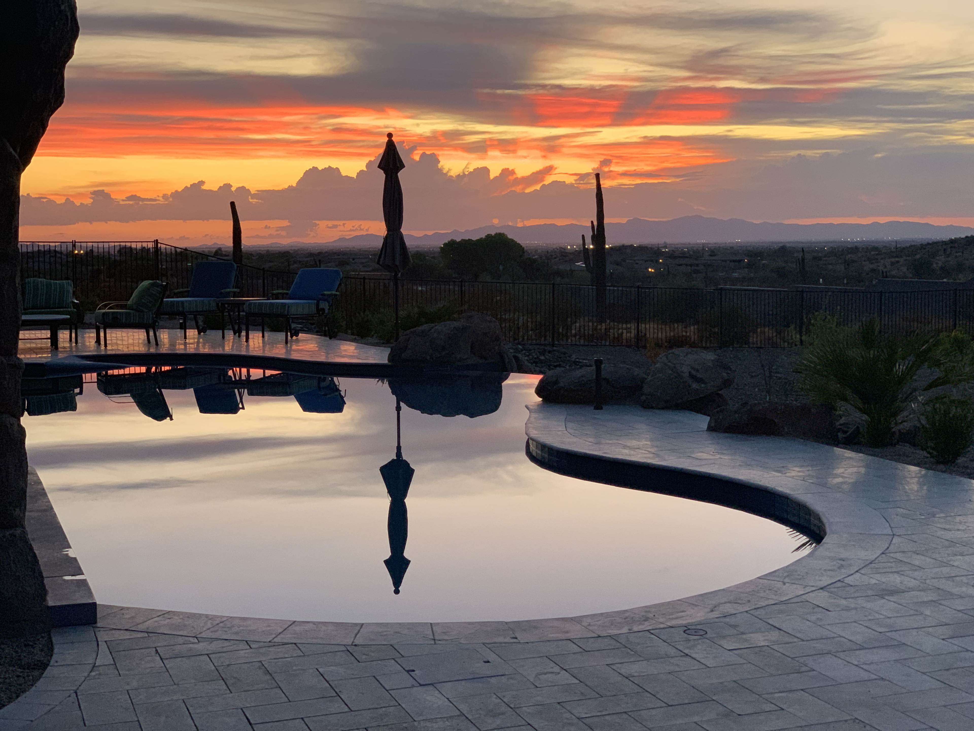 The scene features a swimming pool reflecting the vibrant colors of a sunset over distant mountains, surrounded by outdoor furniture and rocky landscaping.