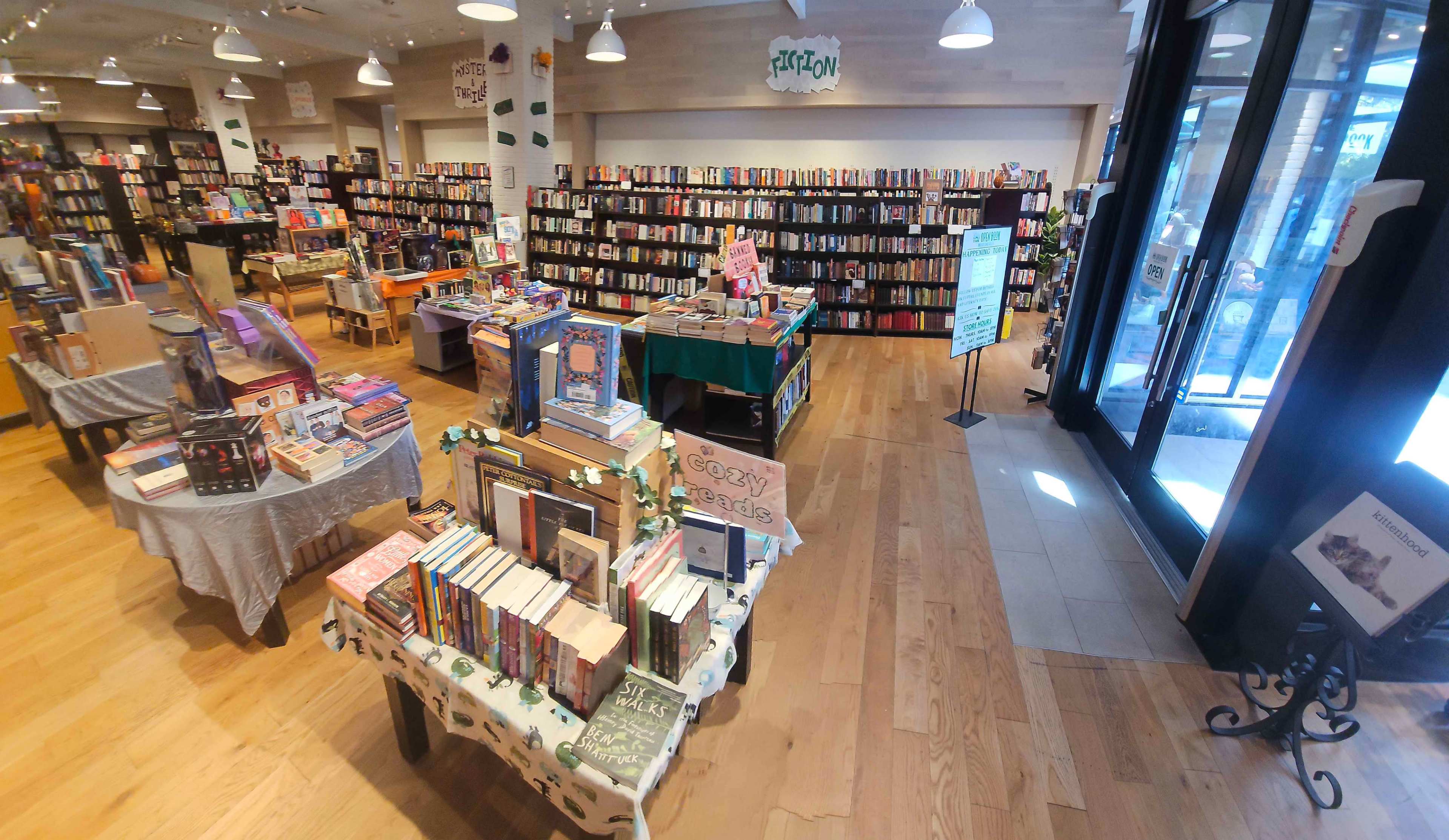 The image shows an interior view of a bookstore with rows of bookshelves filled with various books, display tables in the foreground, and large windows providing natural light.