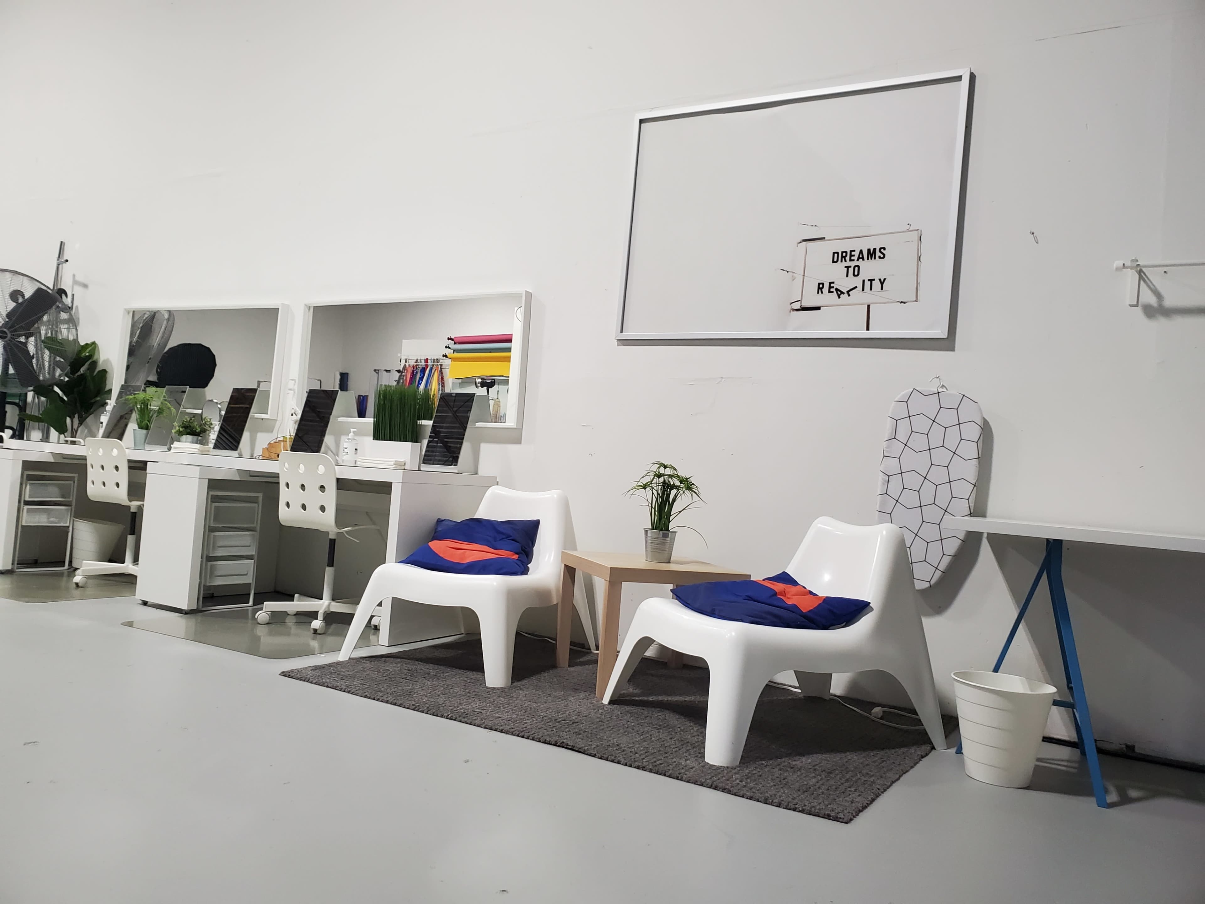 A minimalist office space featuring two white chairs, a small wooden table, potted plants, and a large empty frame on the wall.