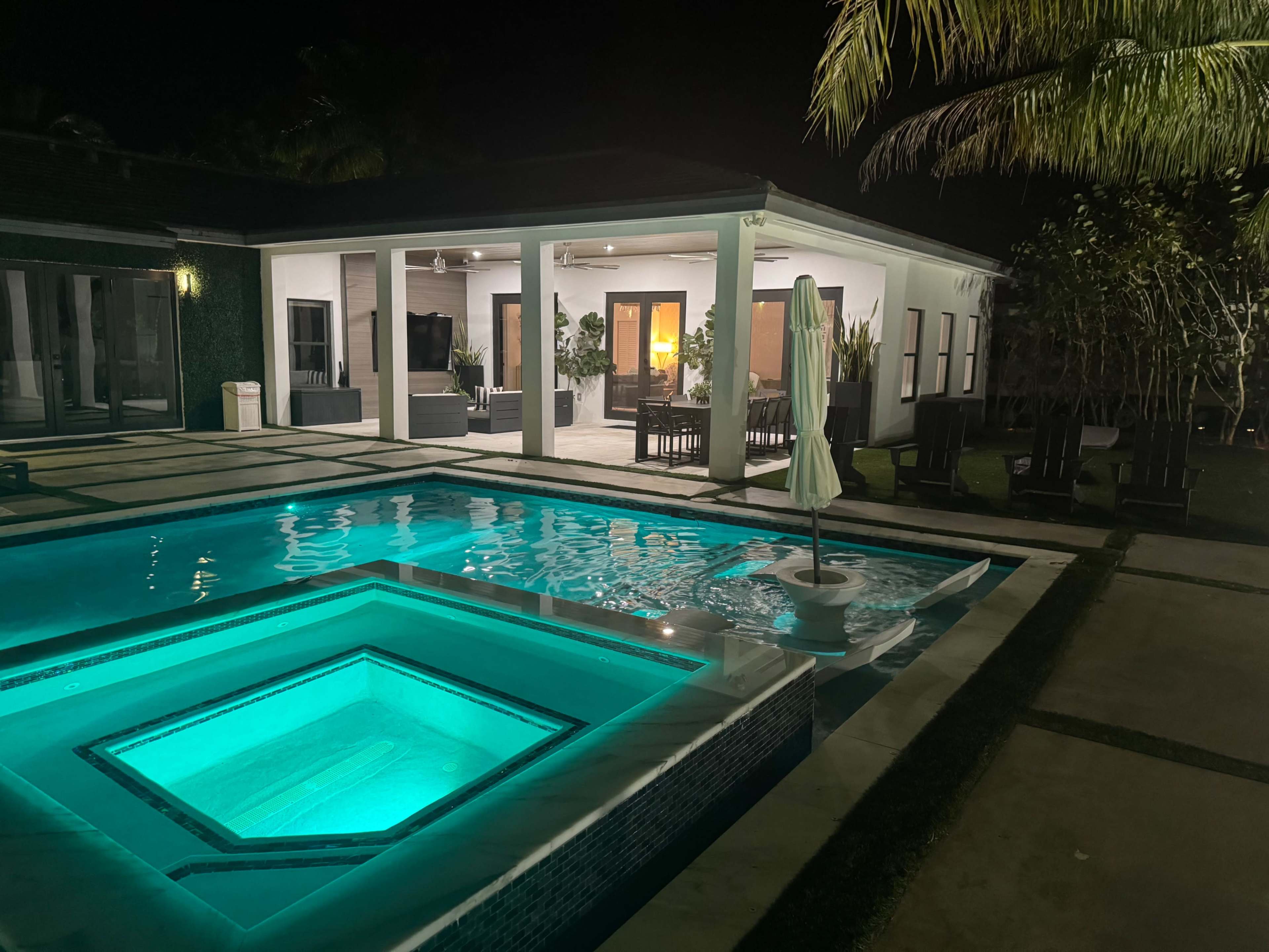 A well-lit swimming pool at night reflects the surrounding landscape and a covered patio area with seating.