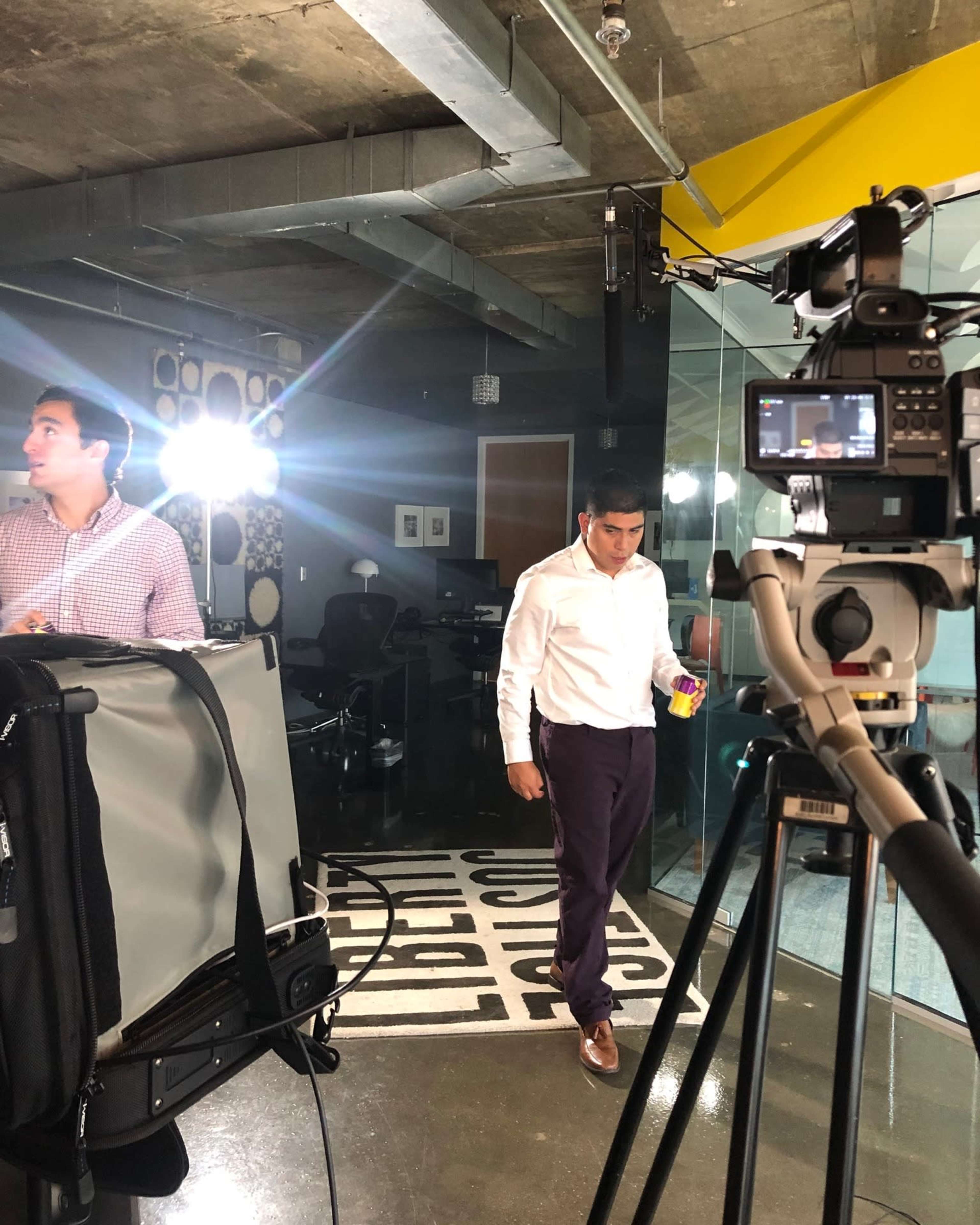 A man in a white shirt walks past a camera setup in an office environment, while another man stands nearby.