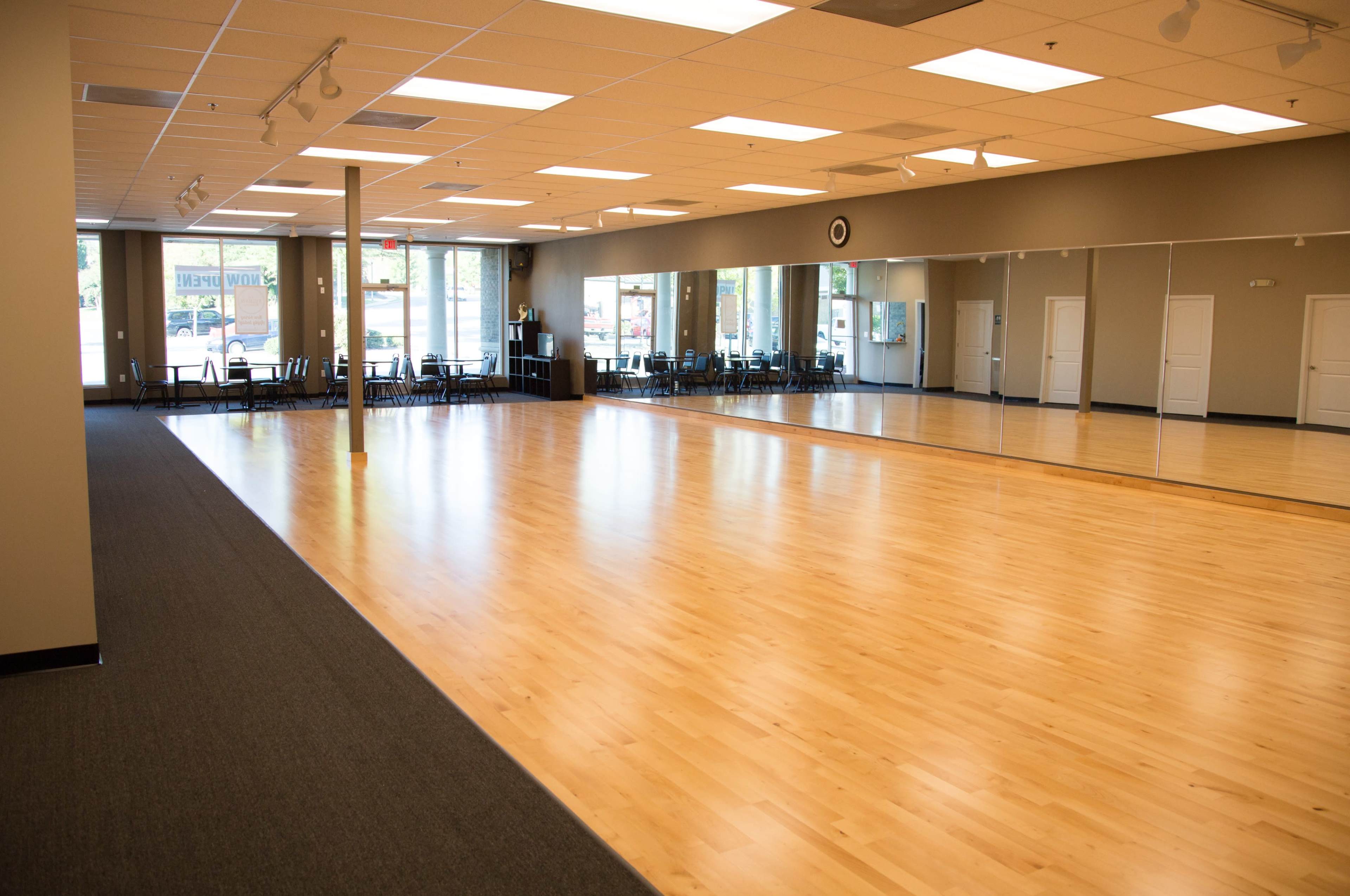 The image shows a spacious dance studio with a wooden floor, large mirrors, and chairs arranged along the sides.