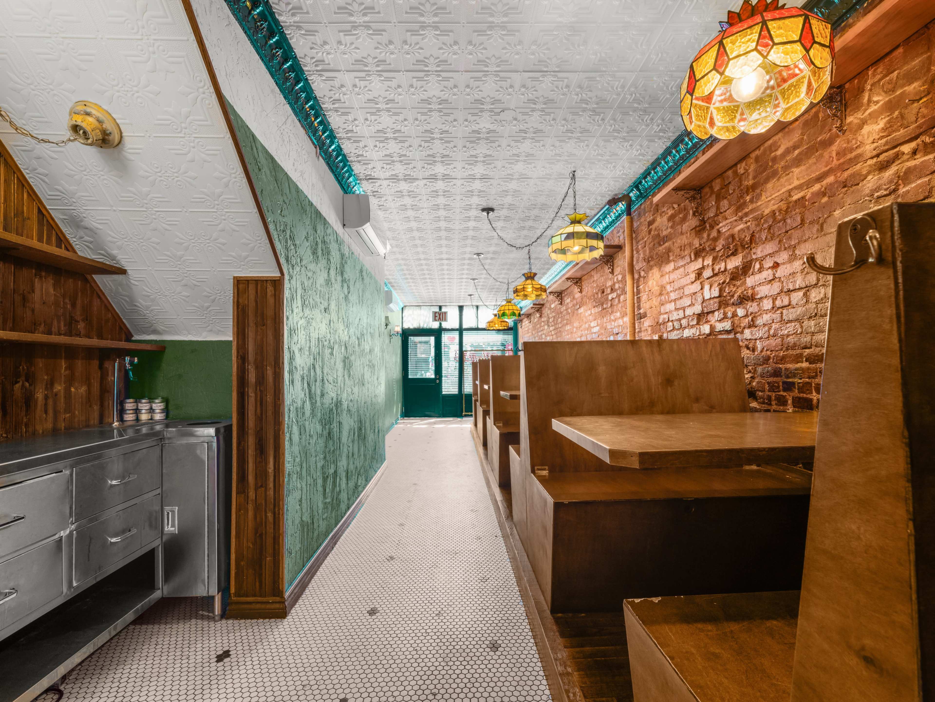 The image shows a narrow, brick-walled restaurant interior with wooden booths, a tiled floor, and vintage-style lamps hanging from the ceiling.