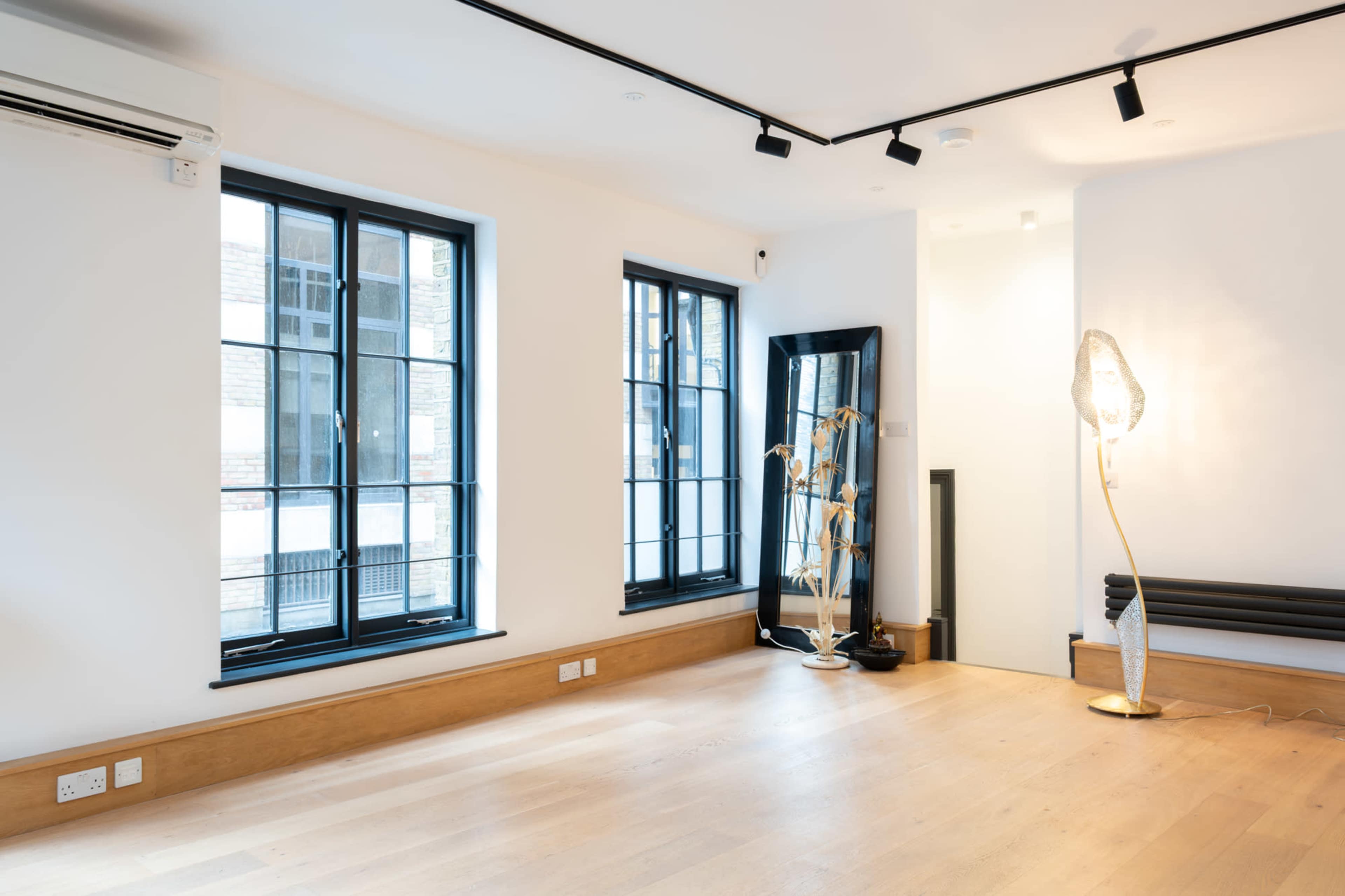 The image shows a modern room with large windows, wooden flooring, and minimal furniture, including a floor lamp and a mirror.