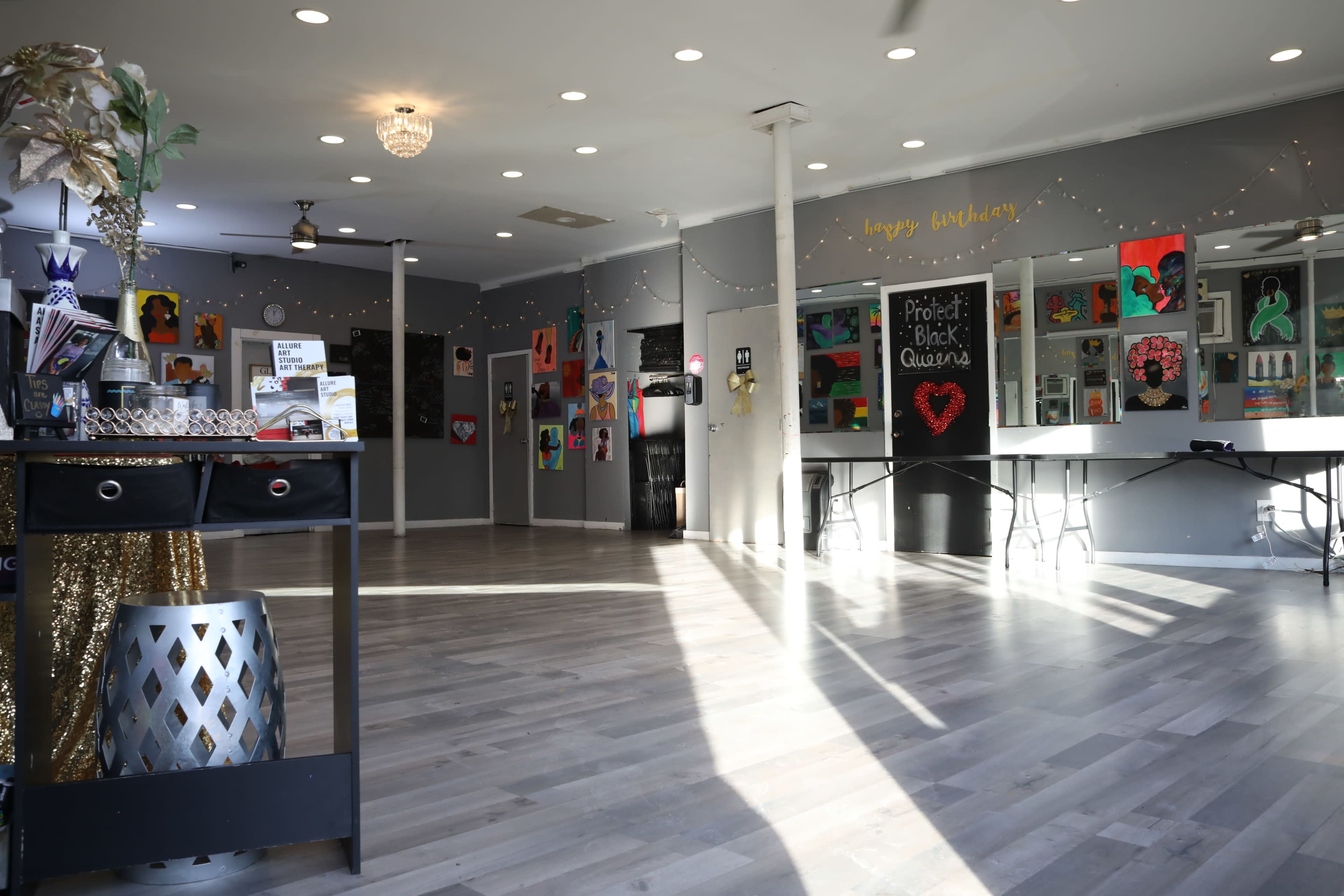 The interior of a gallery space features colorful artwork displayed on the walls, with tables set up for an event and natural light streaming through large windows.