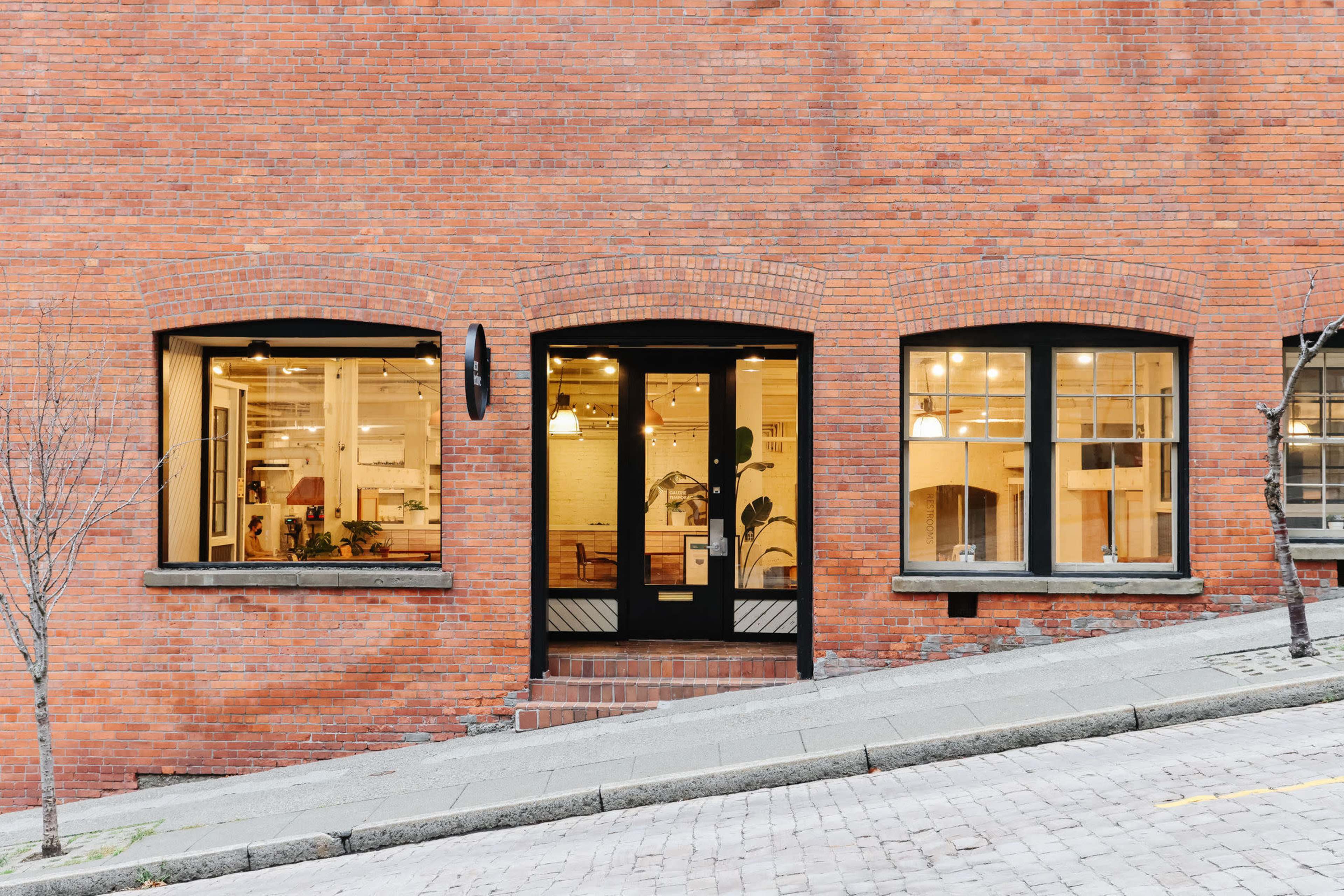 A brick building with large windows and a black door, located on a sloped street.