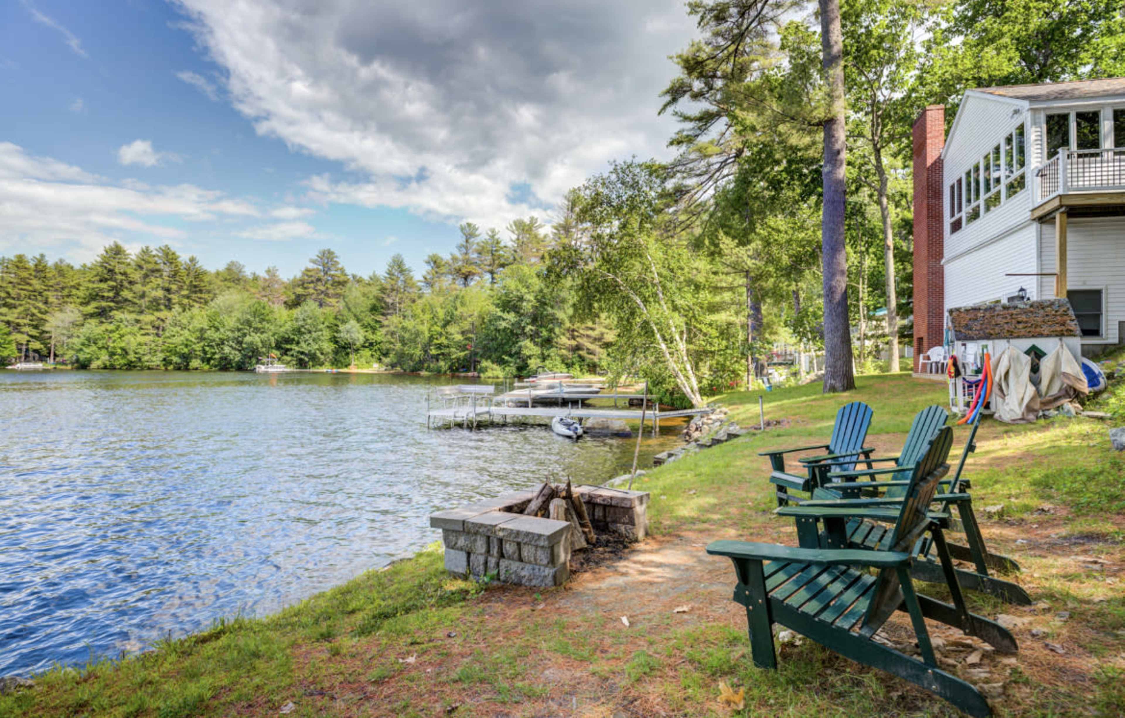 A view of a lakeside area featuring three green Adirondack chairs near a stone fire pit, with a house and dock visible in the background surrounded by trees.