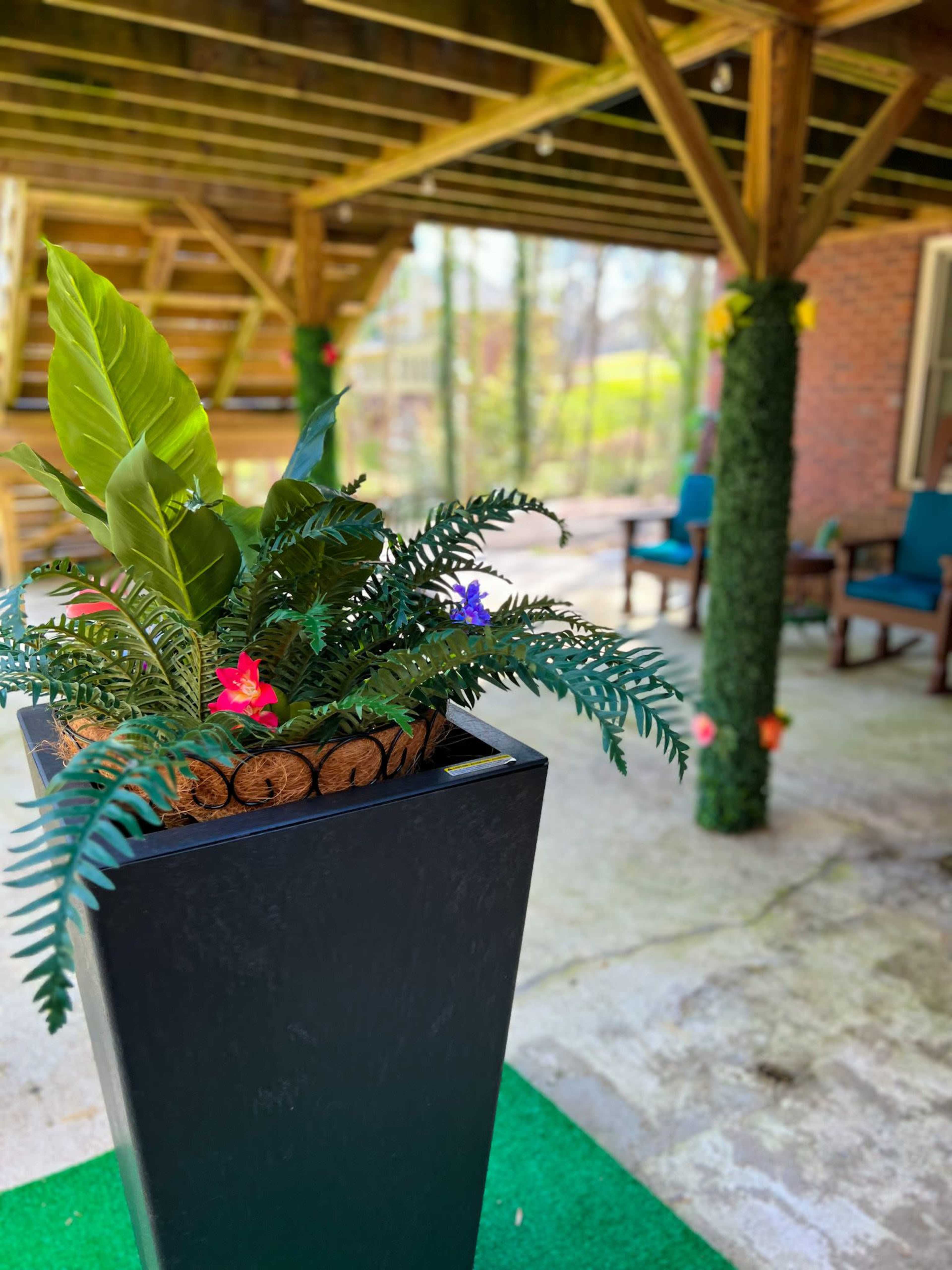 A large planter containing tropical plants is positioned in a covered outdoor area with wooden beams and seating in the background.