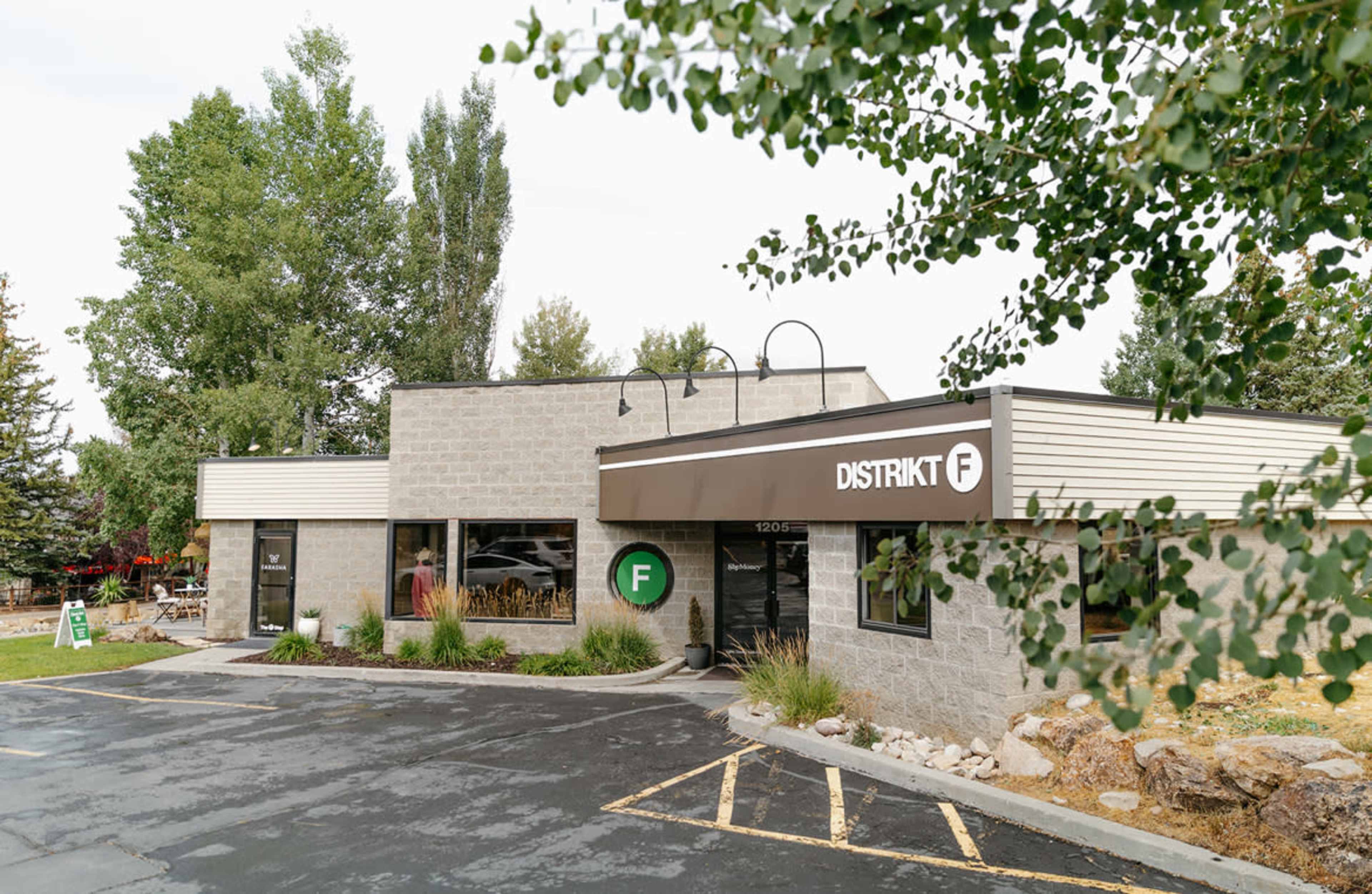 The image shows a modern building with a stone exterior and a large sign that reads "DISTRIKT F," surrounded by greenery and a paved parking lot.
