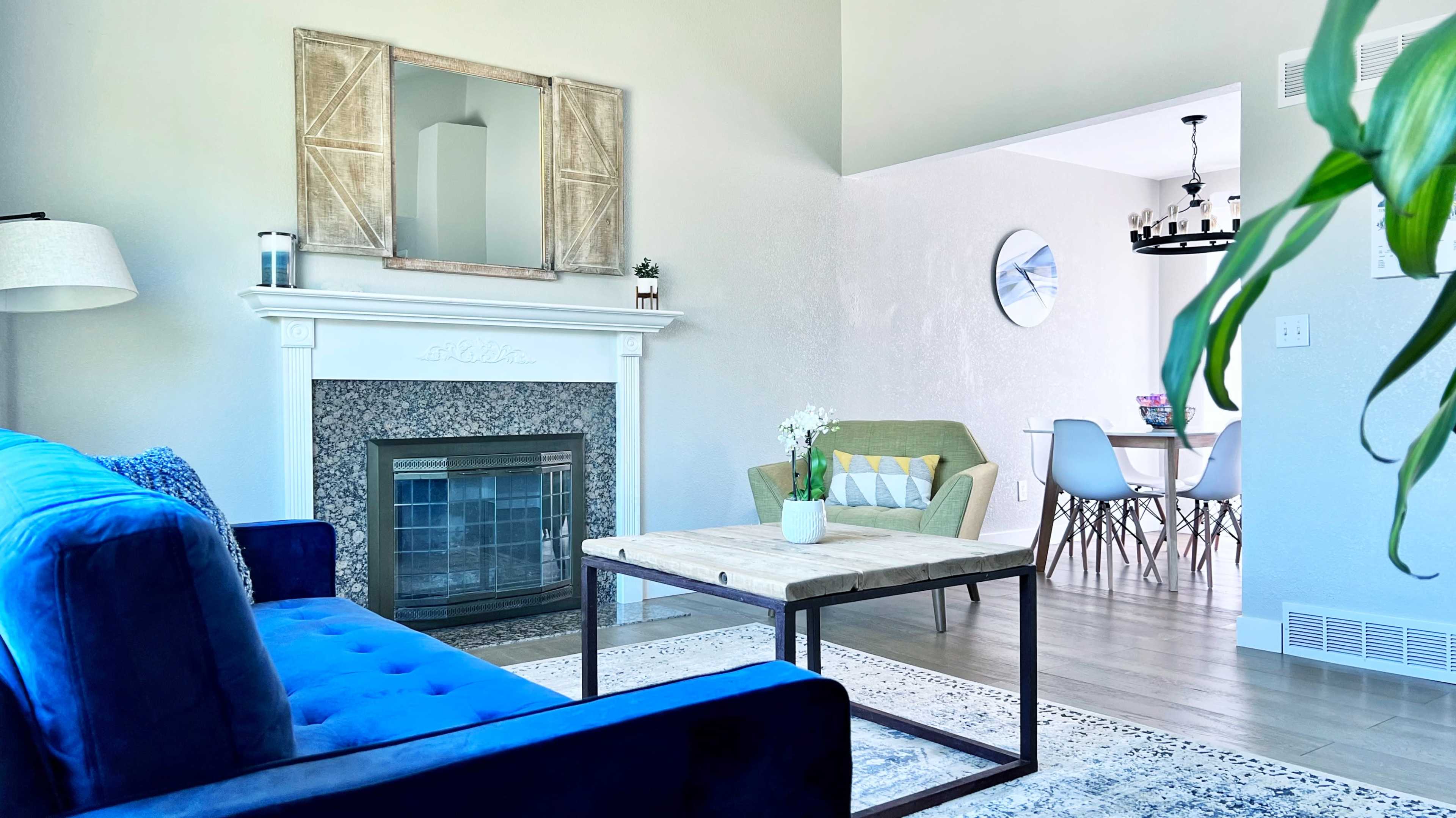 A bright living room features a blue couch, a beige armchair, a coffee table, and a fireplace surrounded by a decorative mantle.