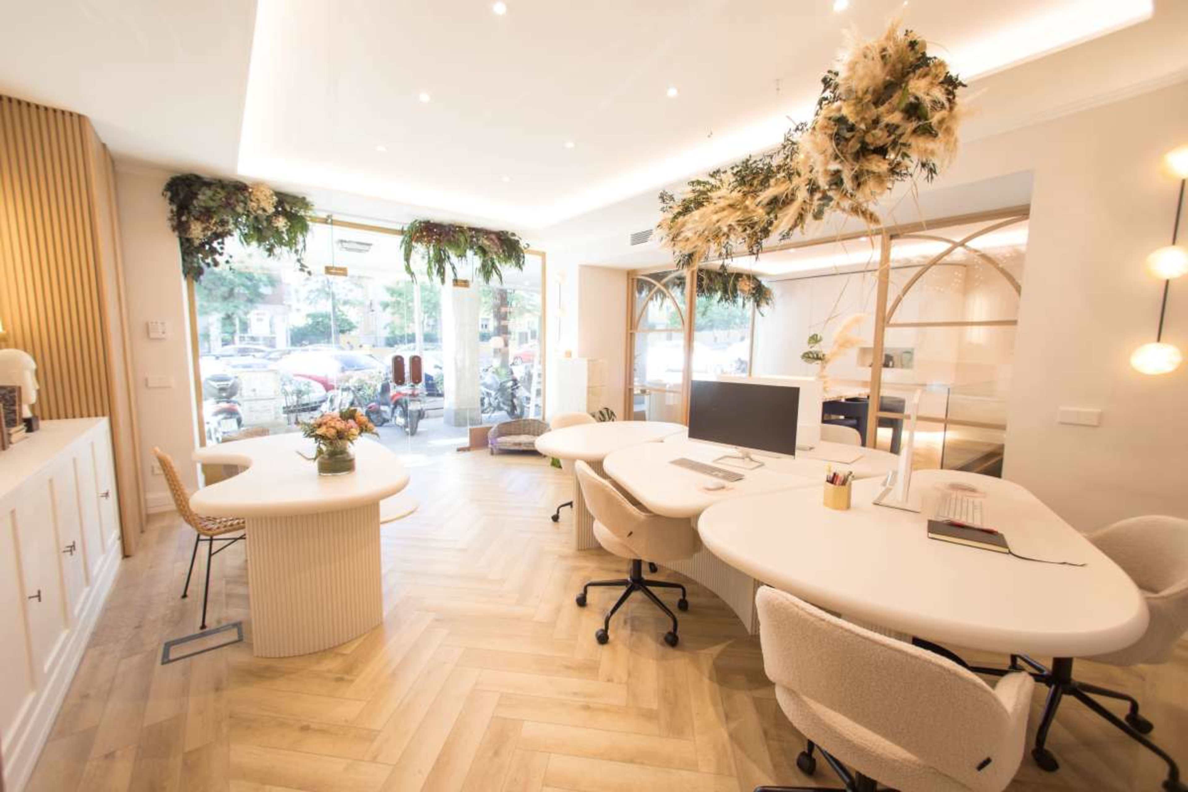The image shows a modern office space with white furniture, wooden accents, and natural light flowing through large windows, decorated with plants and floral arrangements.
