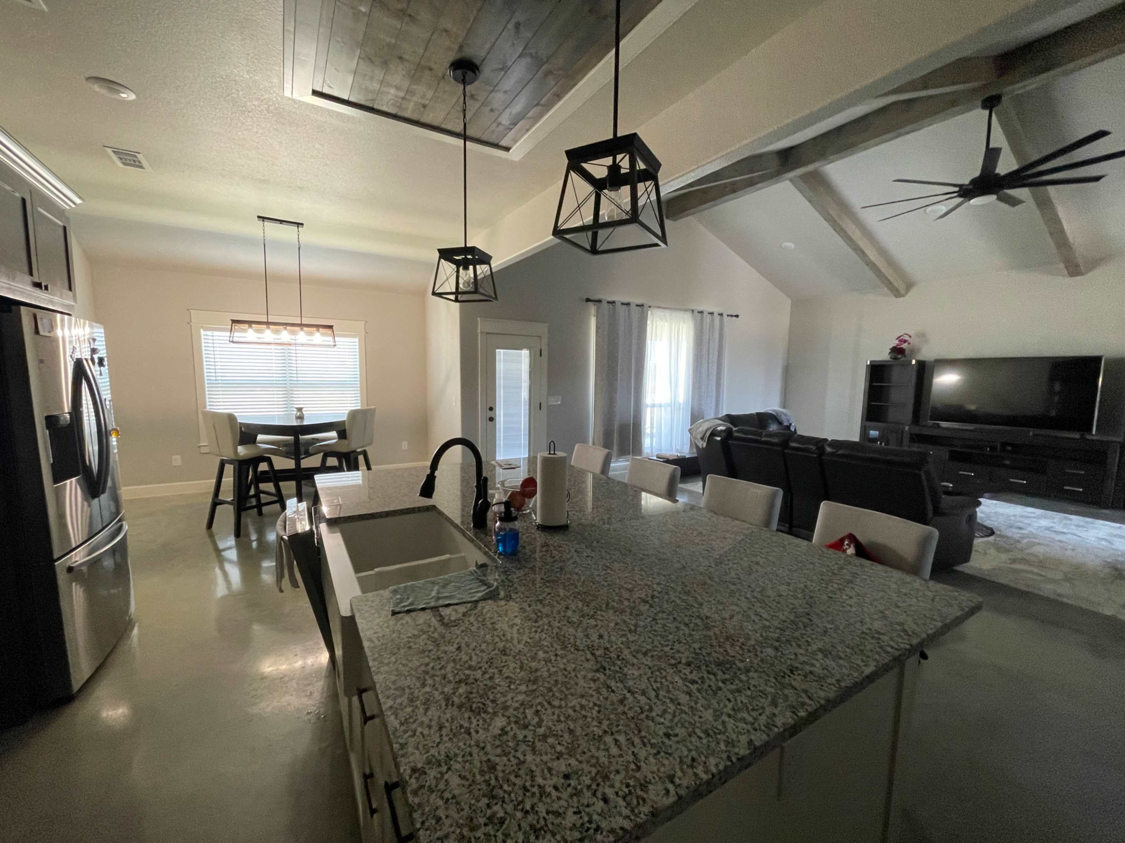 A modern kitchen and living area with a large island, dining table, and a TV stand in a well-lit, open-concept space.