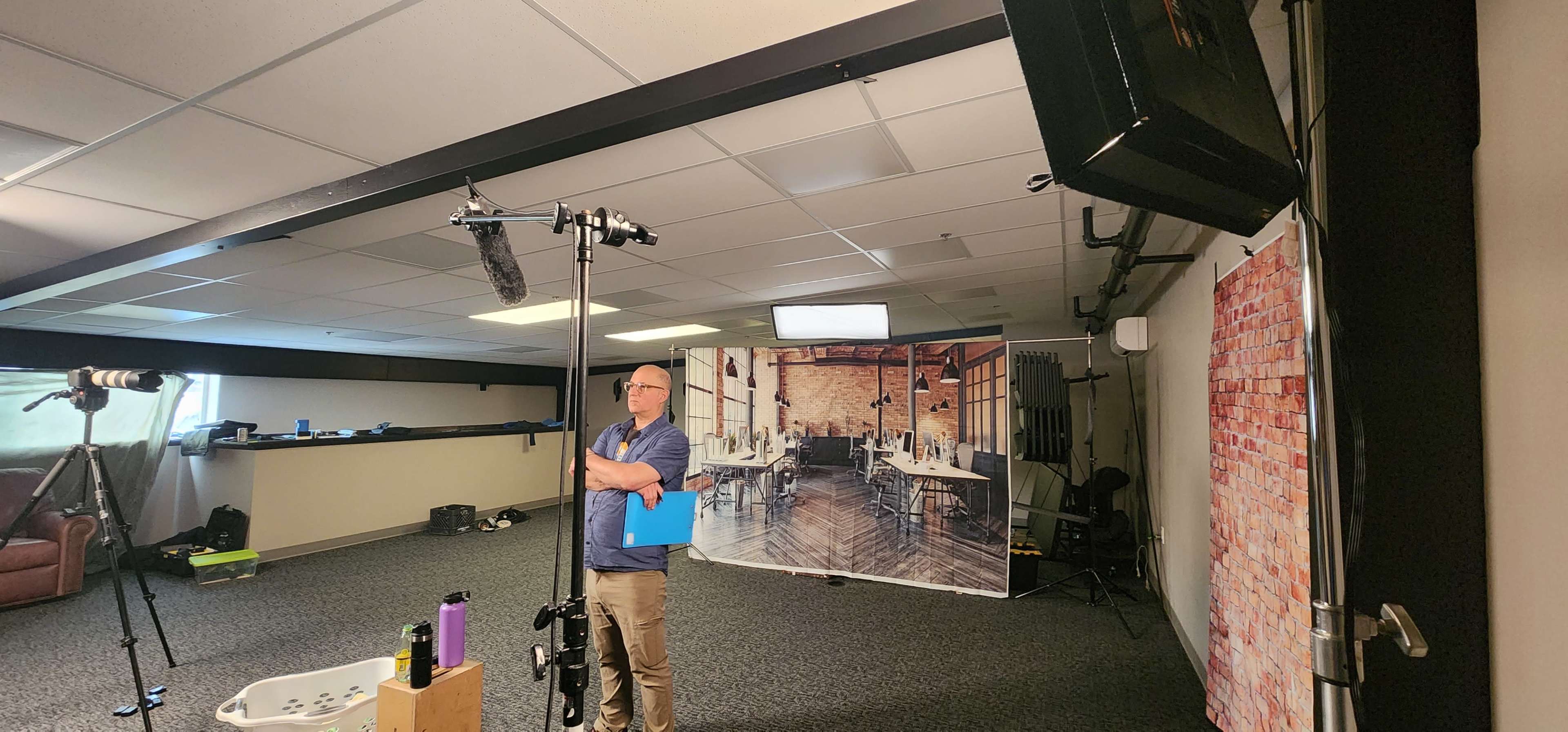 A man stands with a clipboard in a studio space set up for filming, featuring a printed backdrop of a restaurant interior and various equipment scattered around.