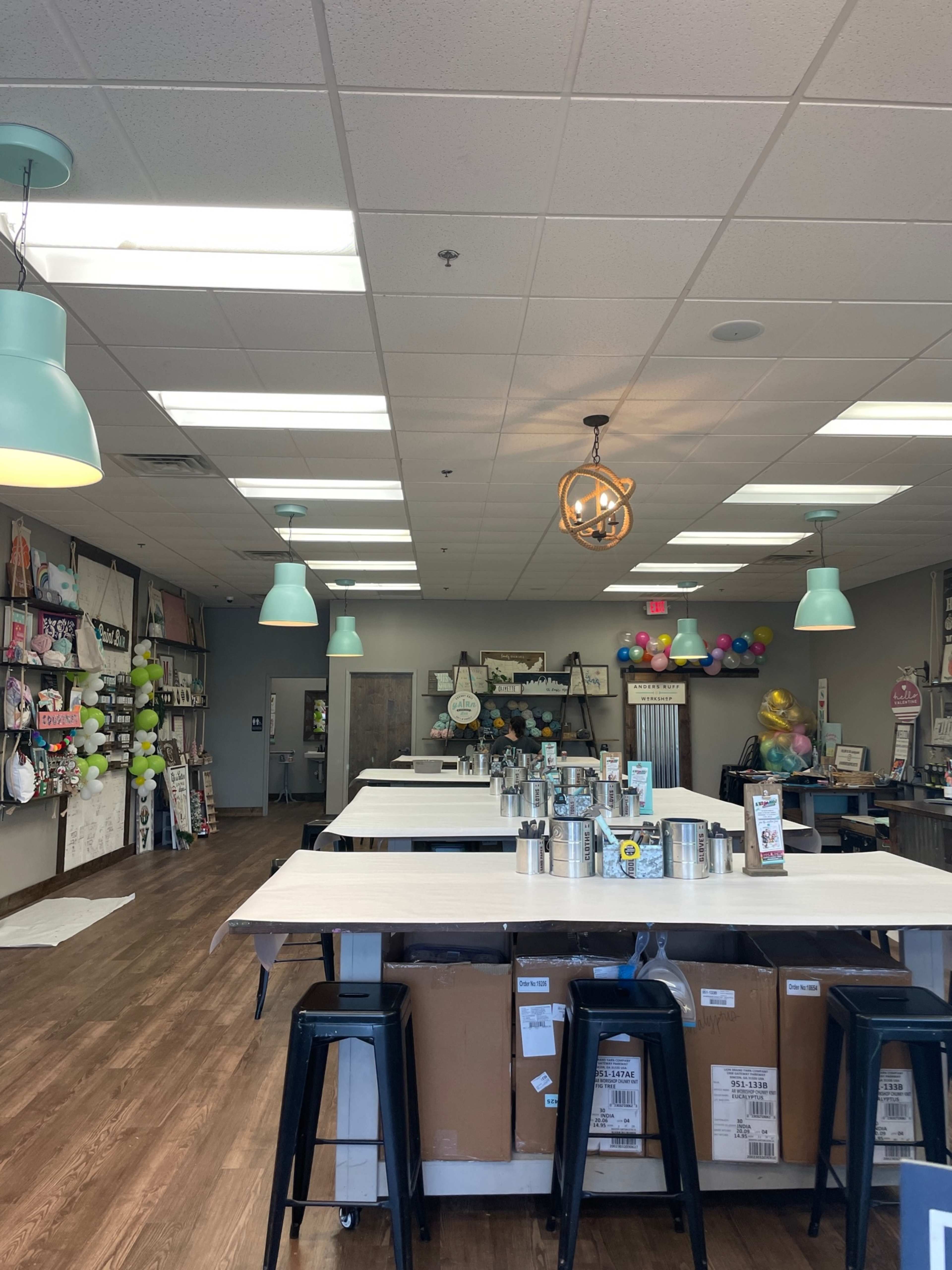 The image shows a spacious, well-lit craft studio with multiple worktables, colorful decorations on the walls, and pendant lighting.