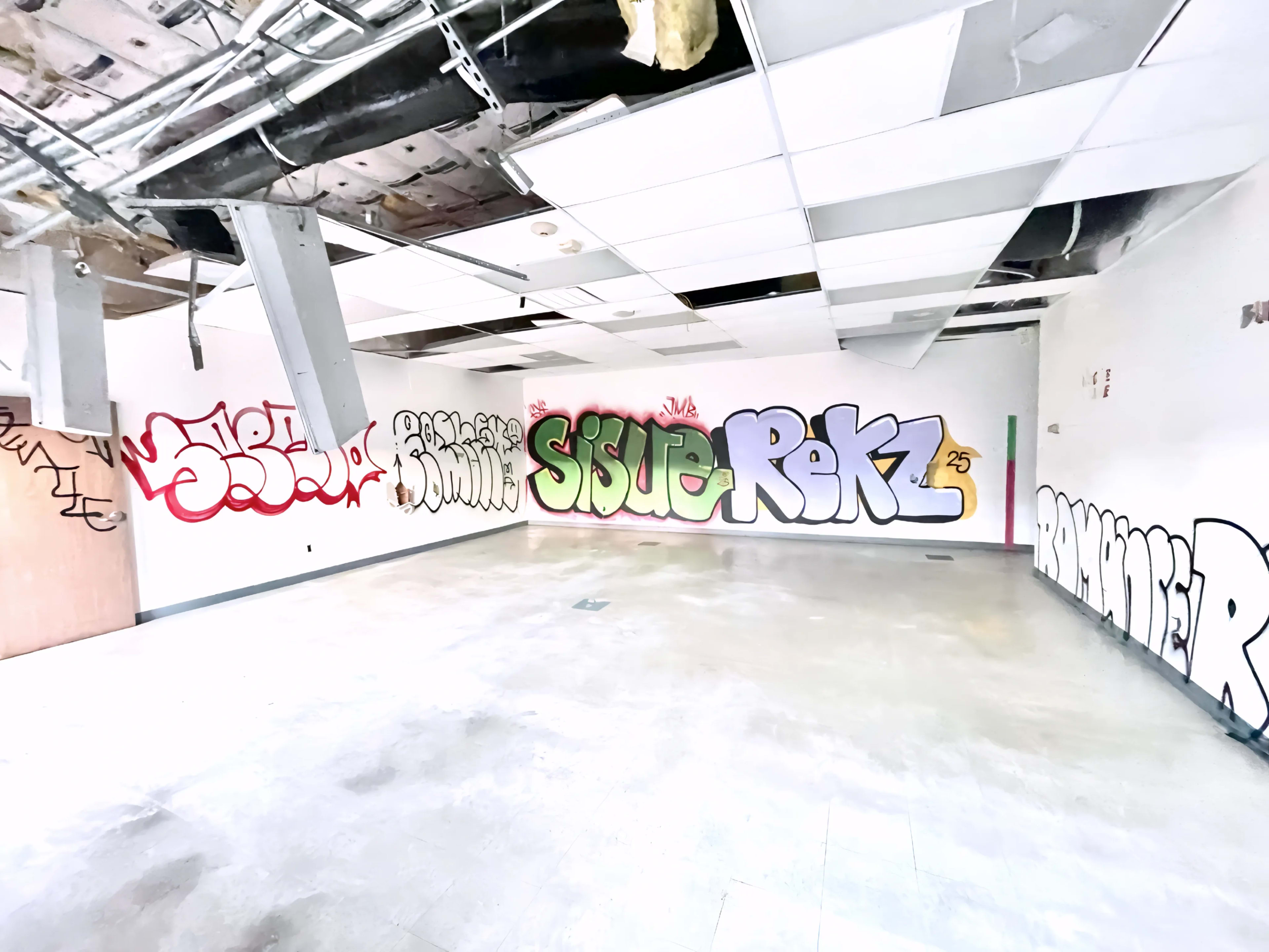 The image depicts an empty room with graffiti covering the walls, featuring various colorful tags and letters, and a damaged ceiling with exposed pipes.