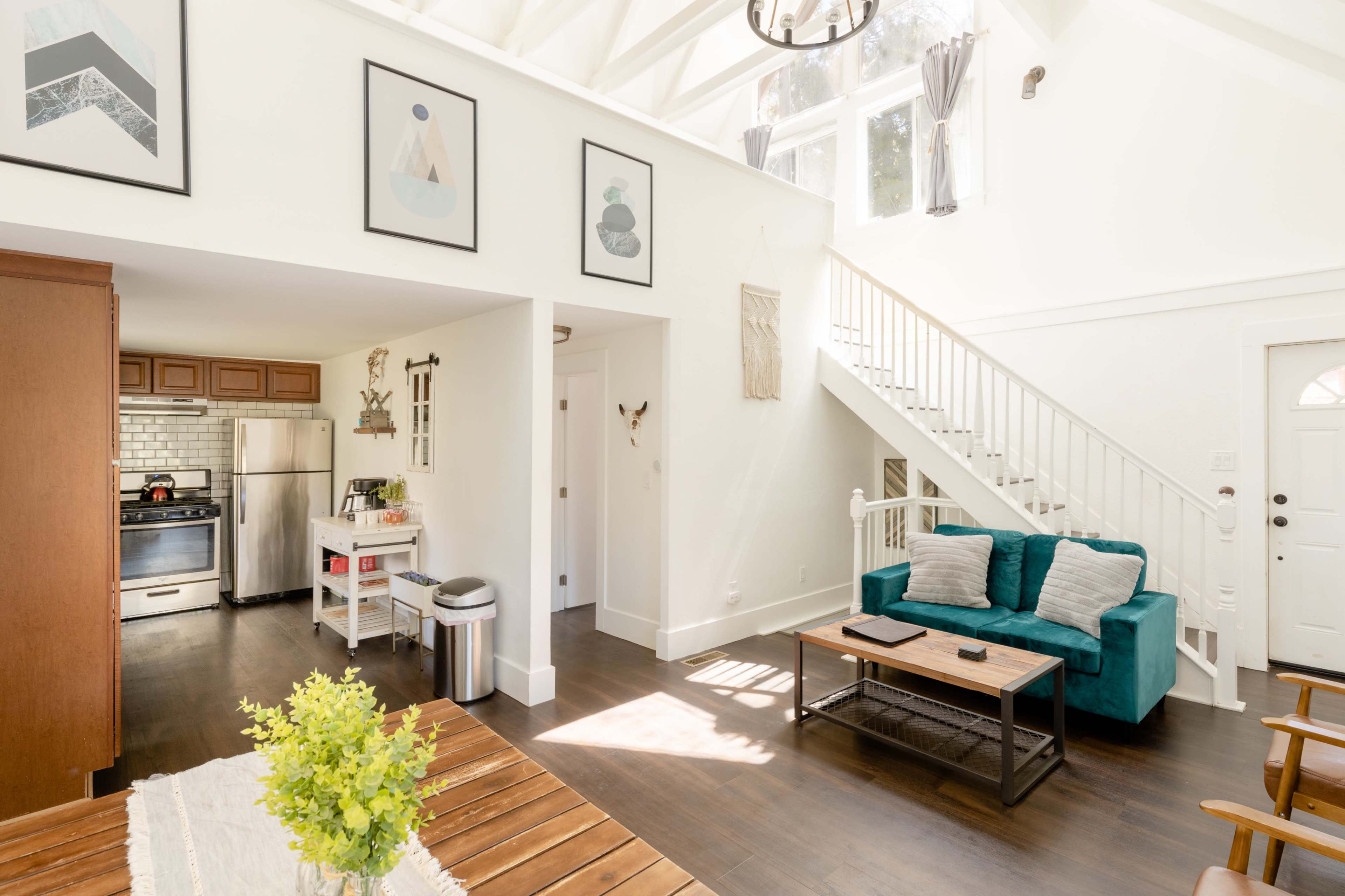 The image depicts a bright, open-concept living space featuring a kitchen area, a staircase, and a seating area with teal couches.