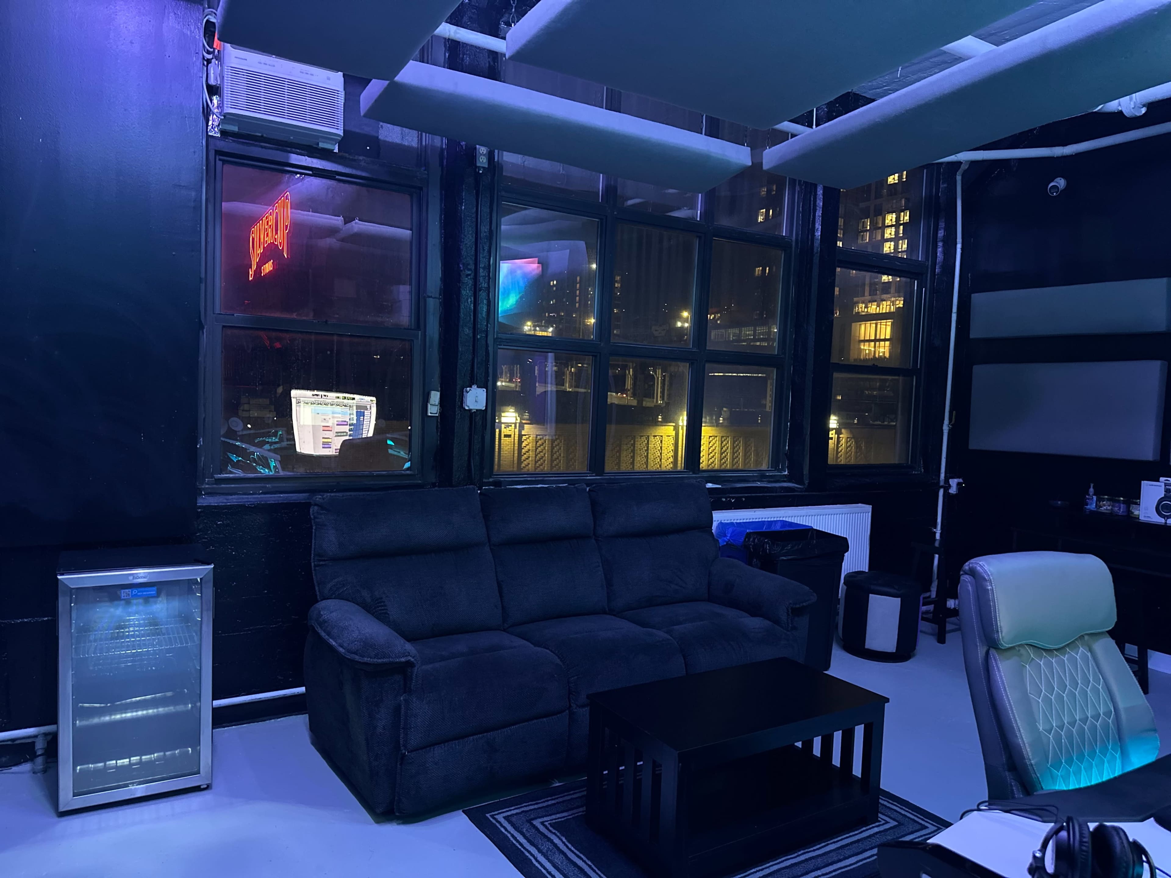 The image shows a dark room with a gray couch, a black coffee table, and a workspace with a chair, illuminated by city lights outside the large windows.