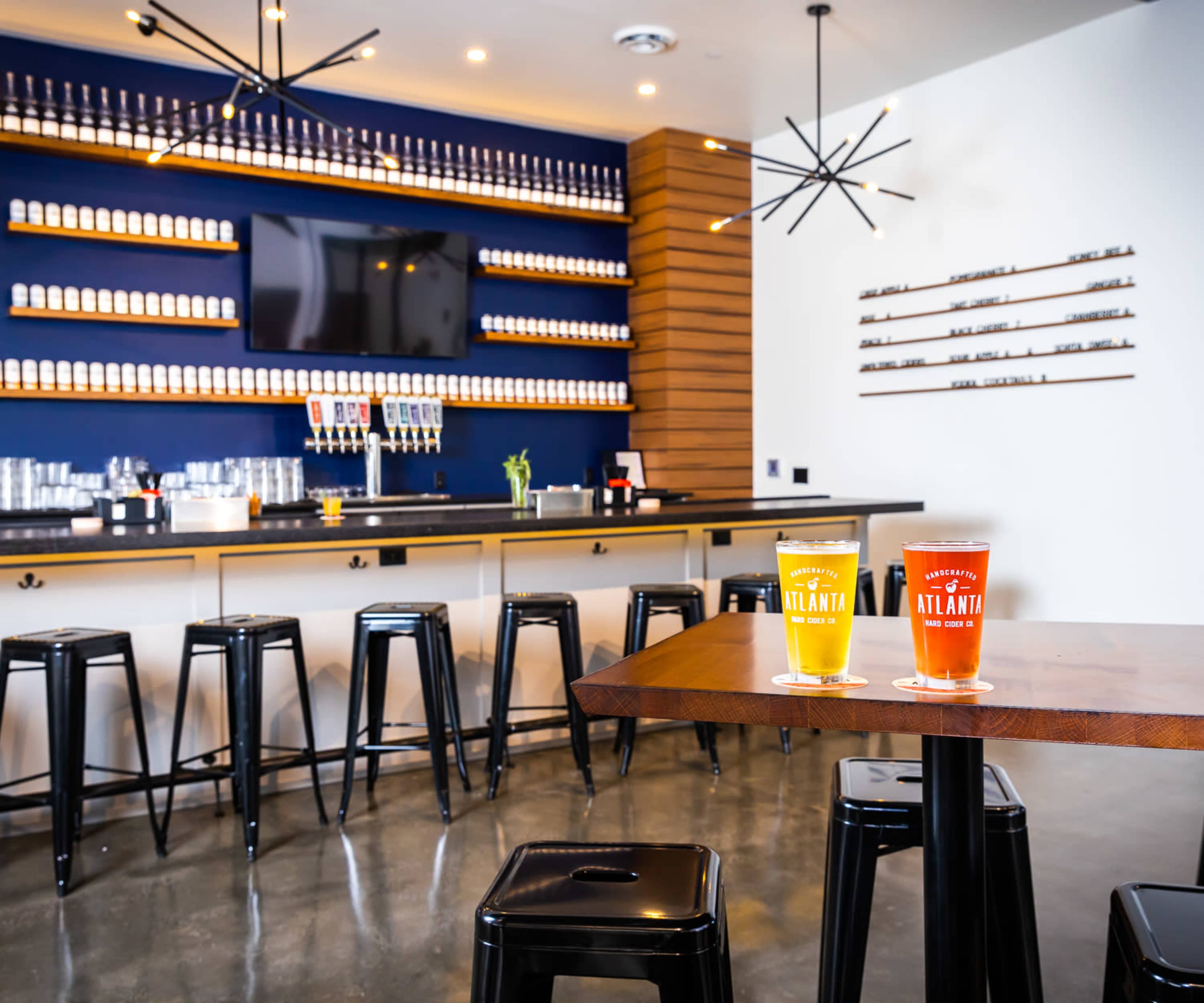 The image shows a modern bar with a display of beer taps and glassware against a wall of beer cans, featuring two glasses of beer on a wooden table.