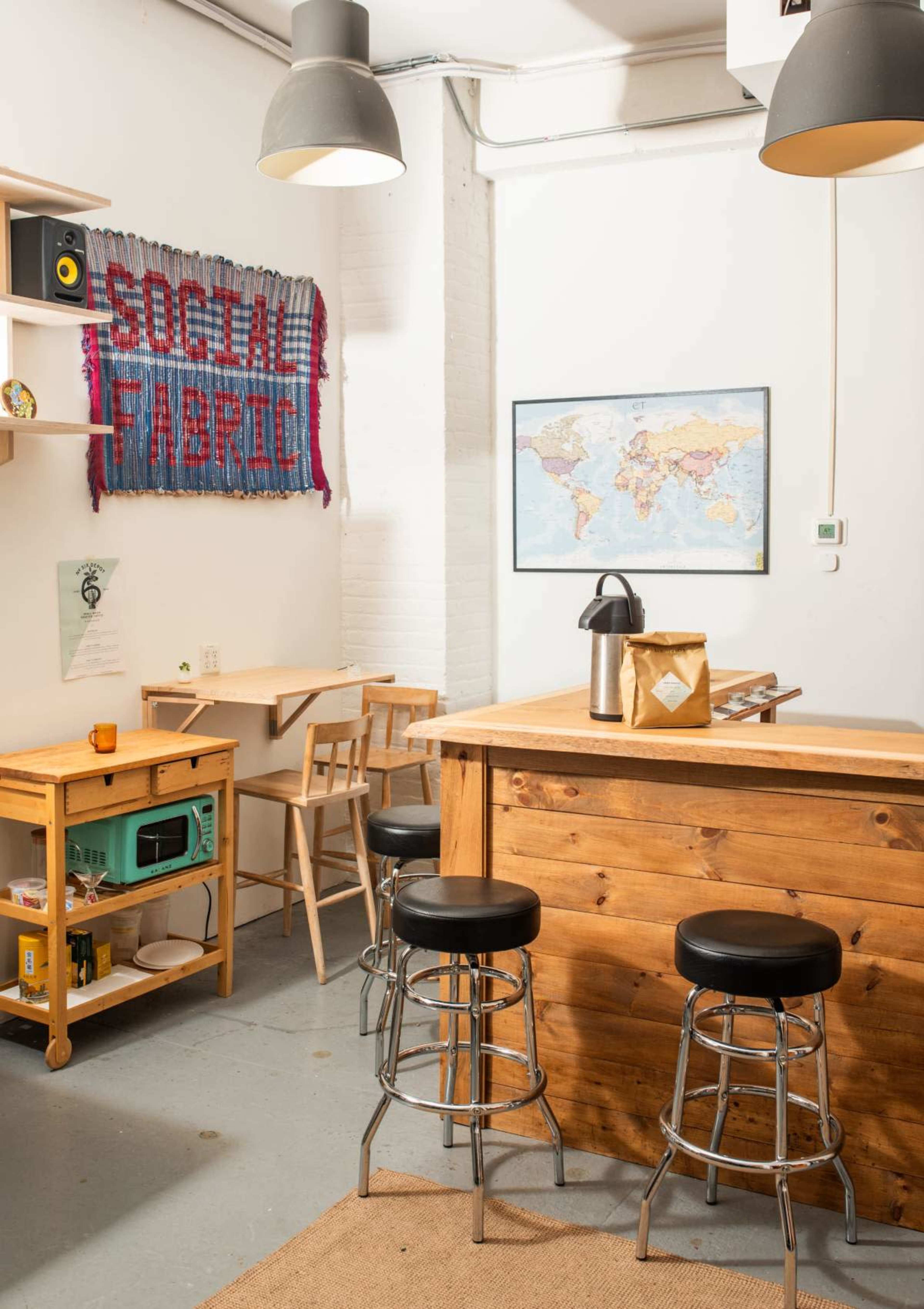 The image shows a bright, minimalistic room with wooden furniture, including a bar counter with stools, a small table, and a wall hanging that says "SOCIAL FABRIC" above a world map.