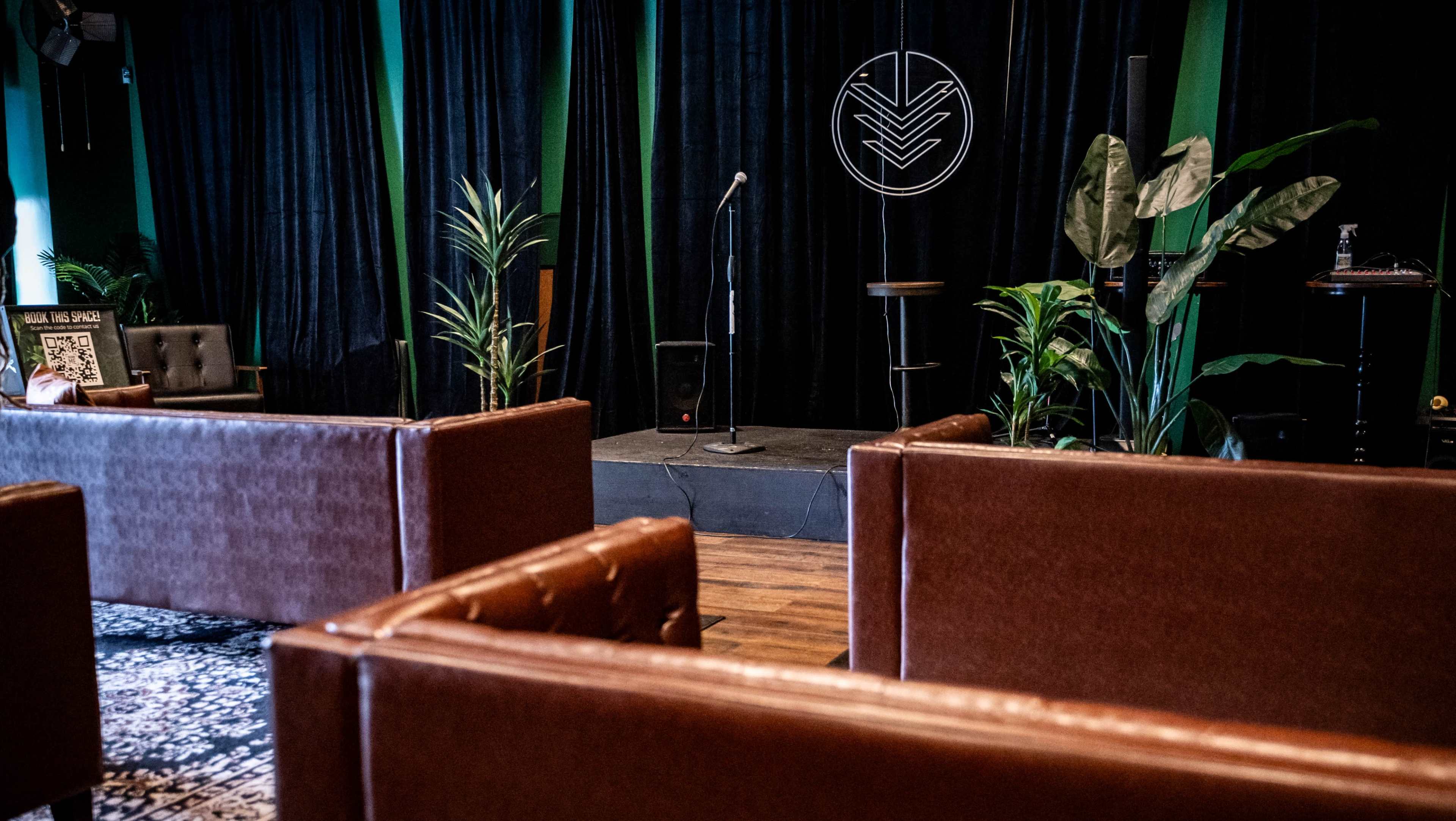 The scene features a small performance stage with a microphone and minimal seating arranged in front, surrounded by potted plants and black curtains.