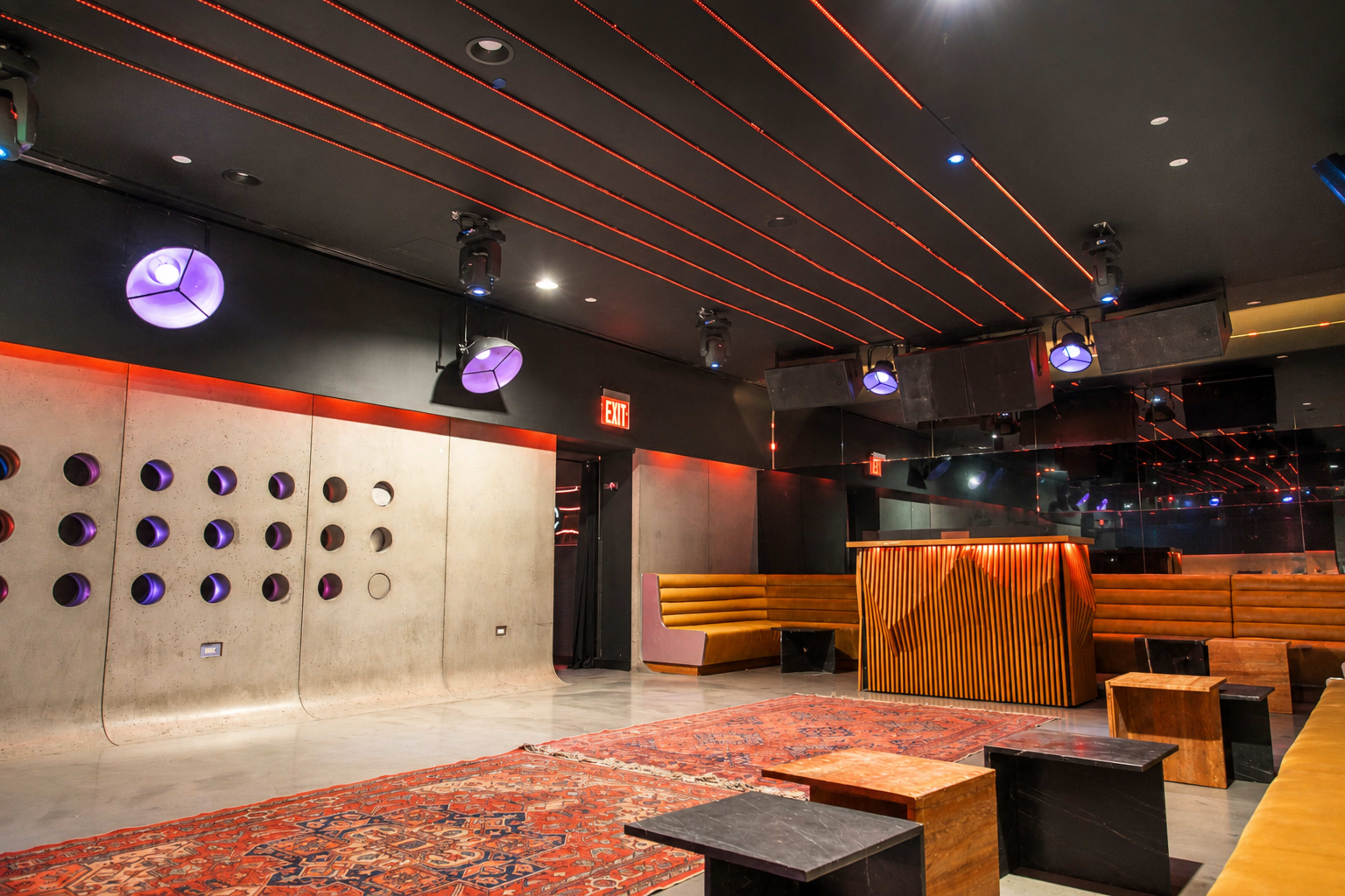 The interior of a modern lounge features a concrete wall with circular cutouts, wooden seating areas, and a patterned rug, illuminated by red and purple lighting.