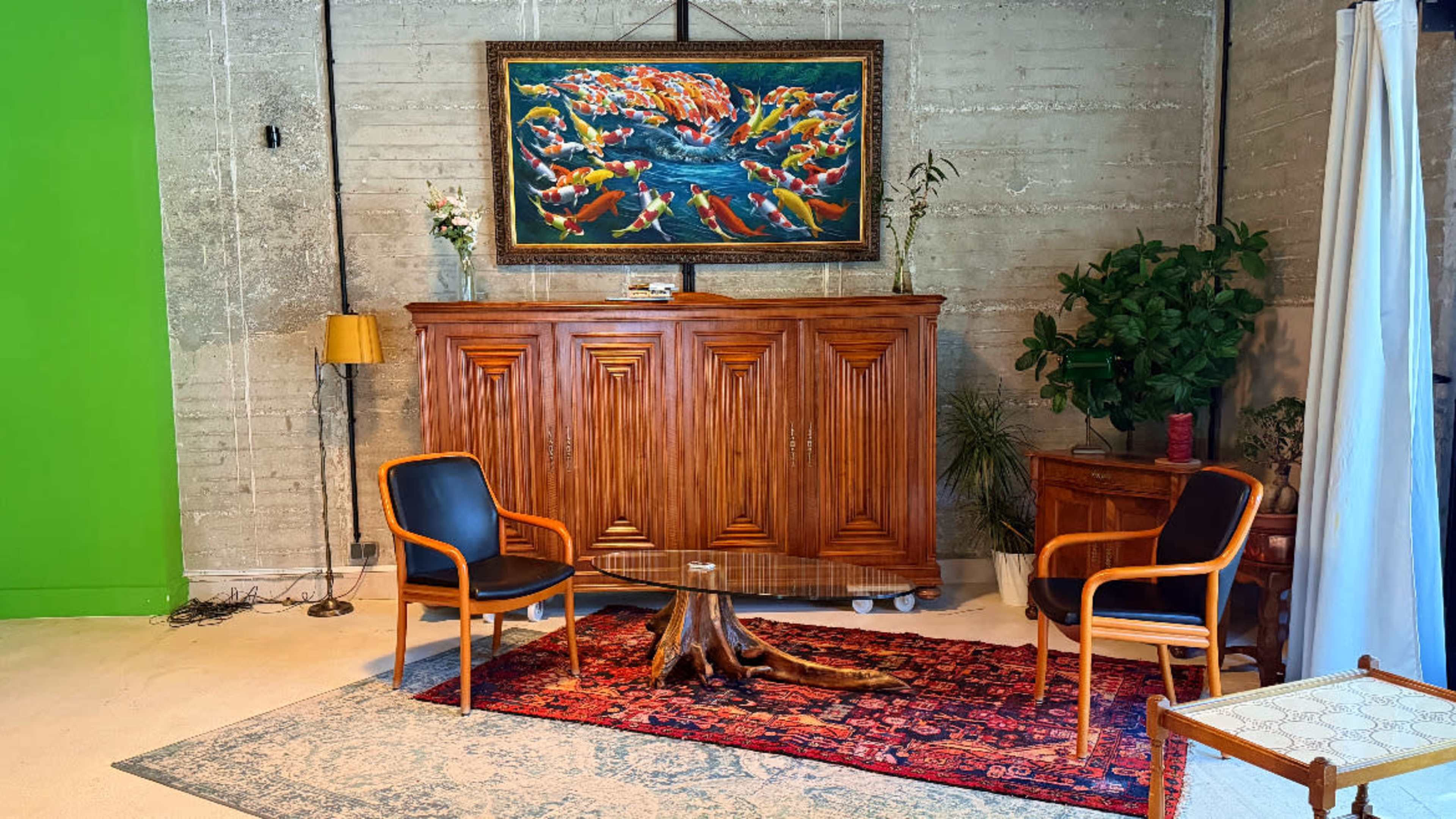 The image shows a room with a wooden cabinet, two black chairs, a colorful painting of fish on the wall, and a potted plant beside a decorative rug.