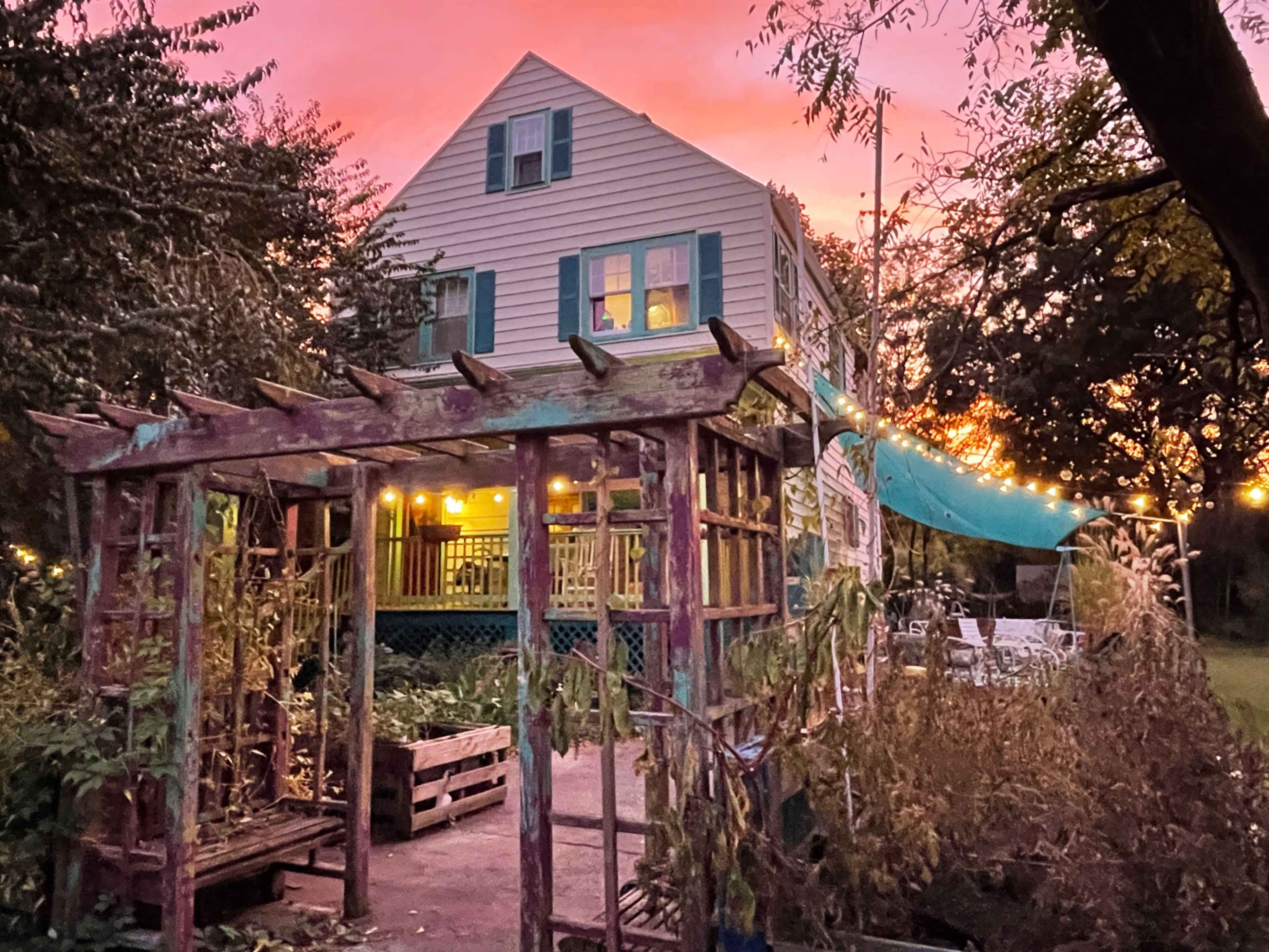 A two-story house with teal shutters is illuminated by evening light under a vibrant pink sky, positioned next to a wooden arbor and garden.