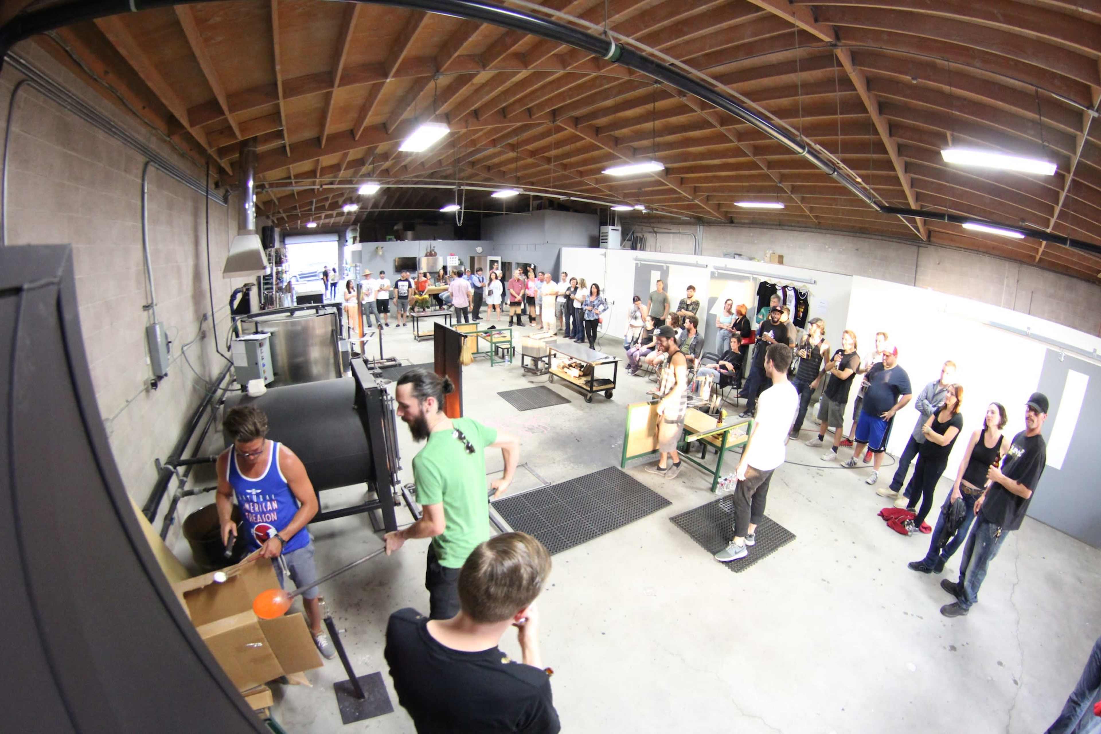 A group of people observes glassblowing demonstrations in an industrial workshop with various equipment and workstations.