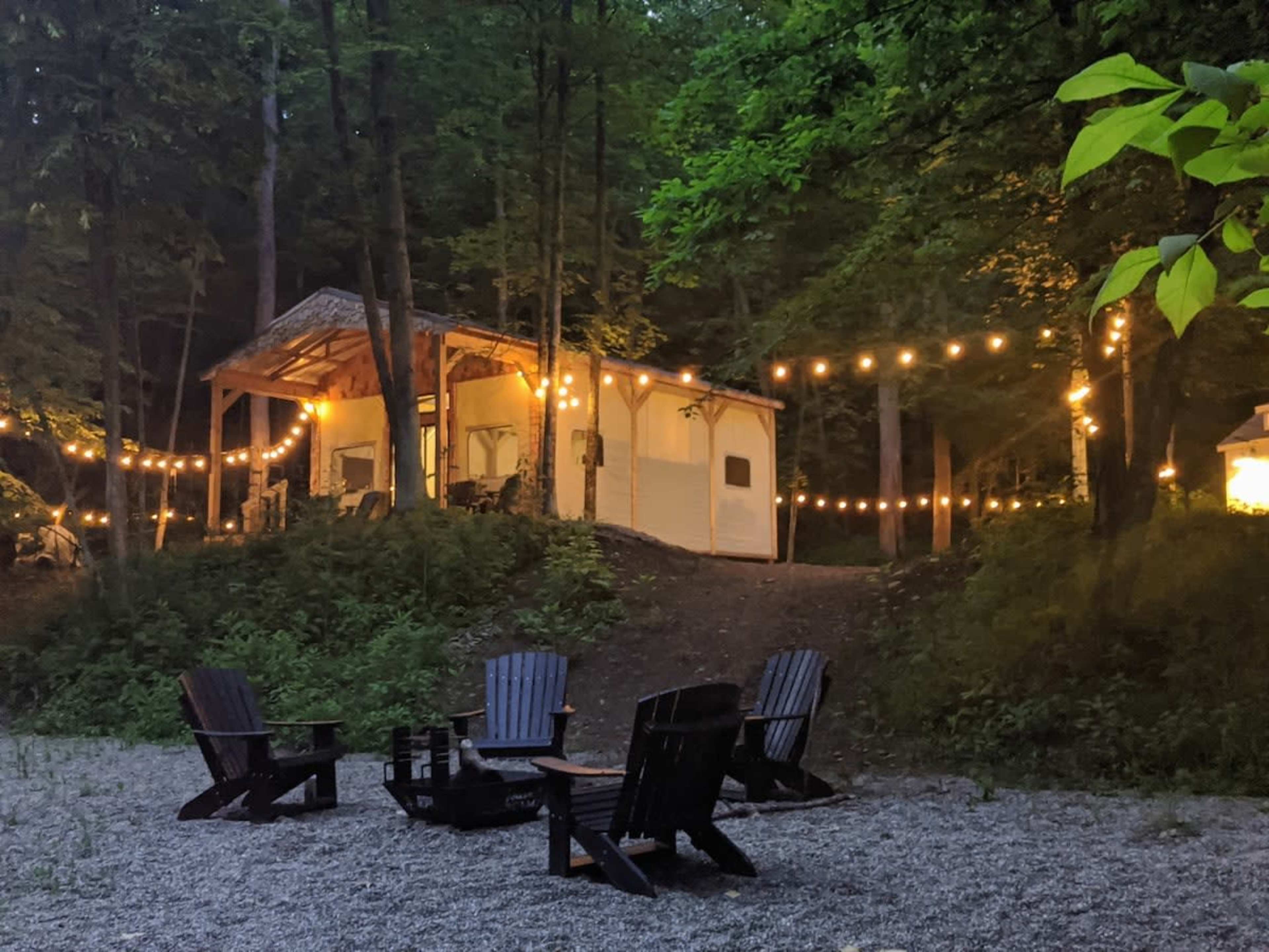 A cabin is illuminated by string lights in a wooded area, with a fire pit surrounded by adirondack chairs in the foreground.