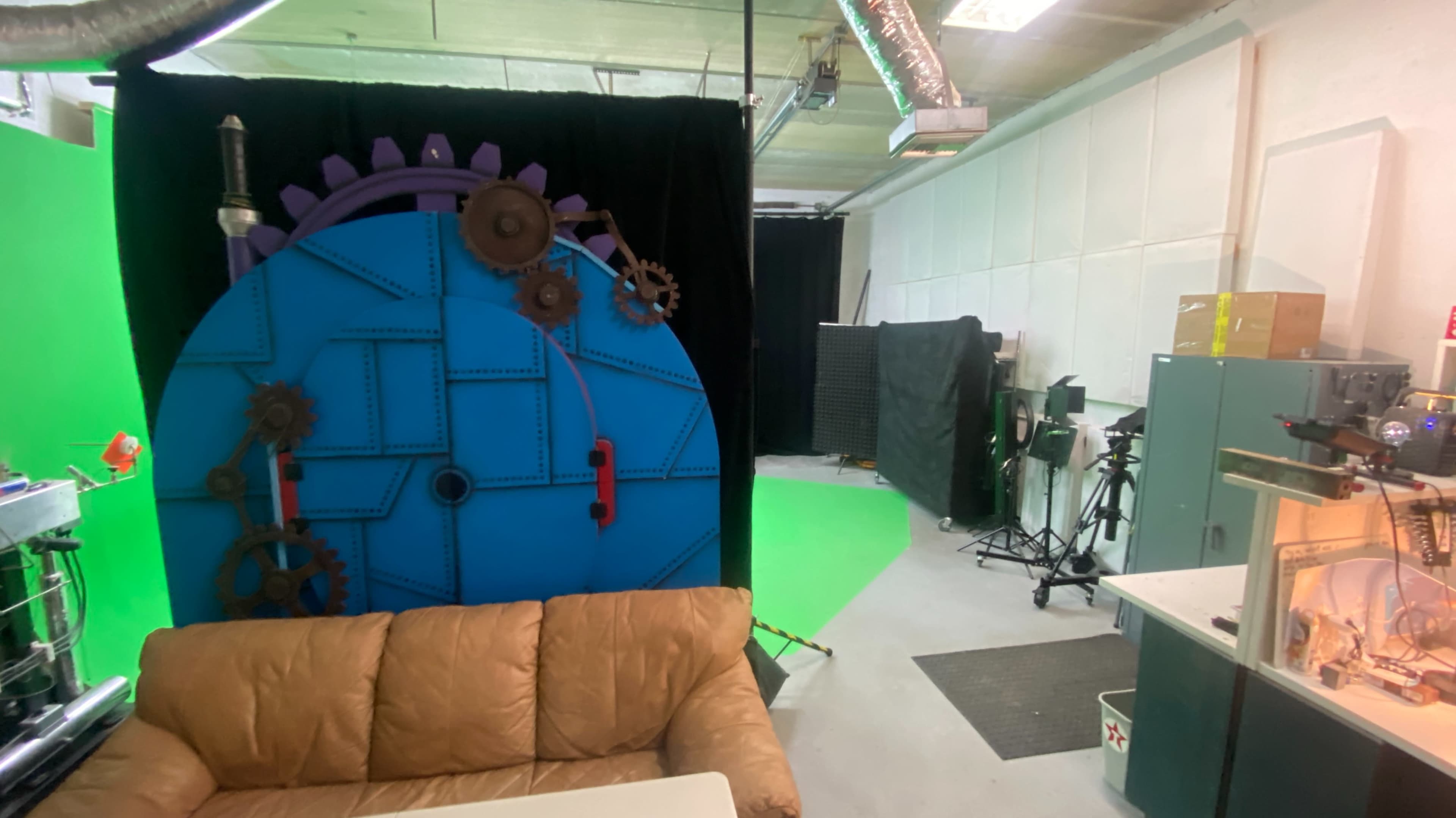 A studio space with a green screen setup, a large blue gear backdrop, a tan couch, and various production equipment in the corners.