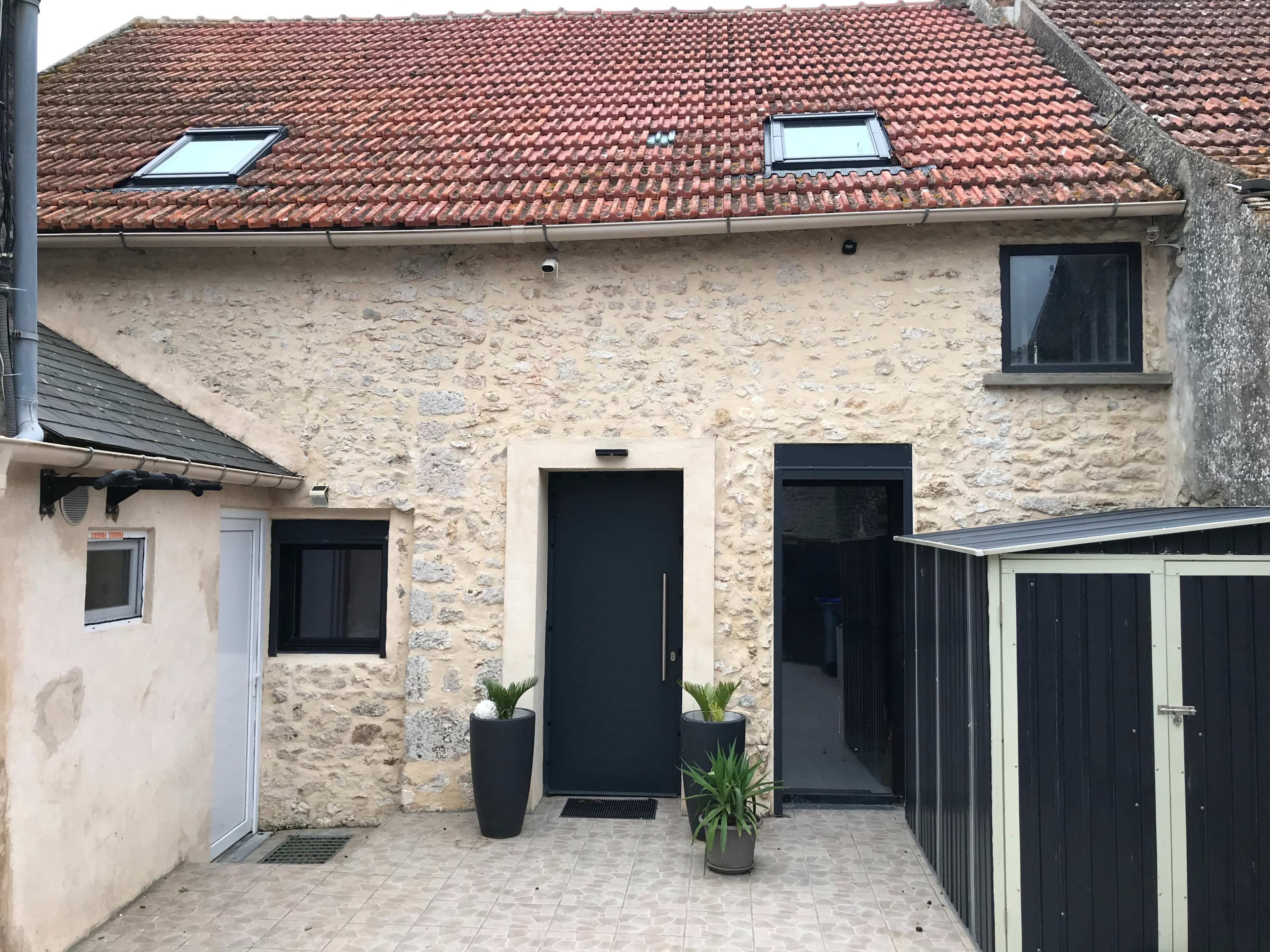 A rustic stone house with a black front door, two windows, and a small patio area with planters.